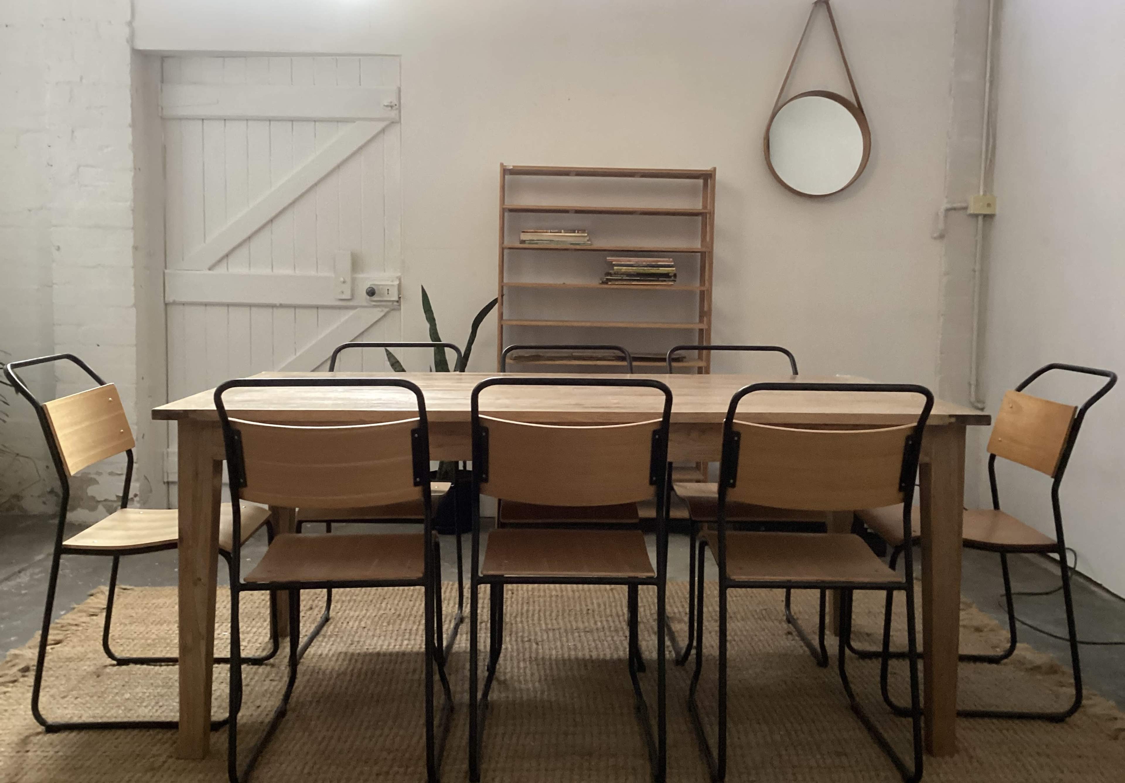 The image shows a wooden dining table surrounded by six metal and wooden chairs in a simple, minimalist room with light-colored walls and a round mirror on the wall.