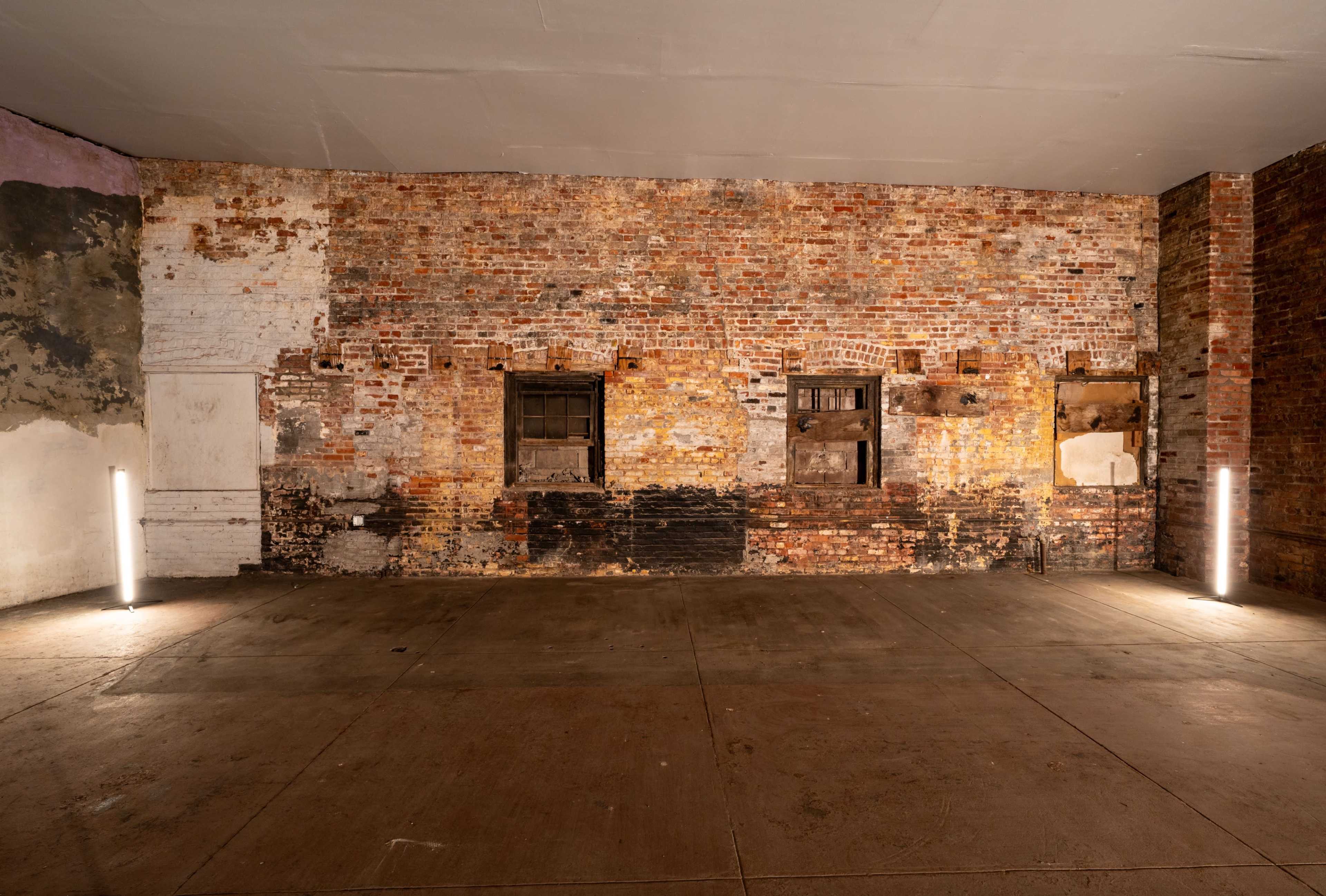 The image shows a spacious, empty room with a textured brick wall featuring several windows and illuminated by two standing lights.