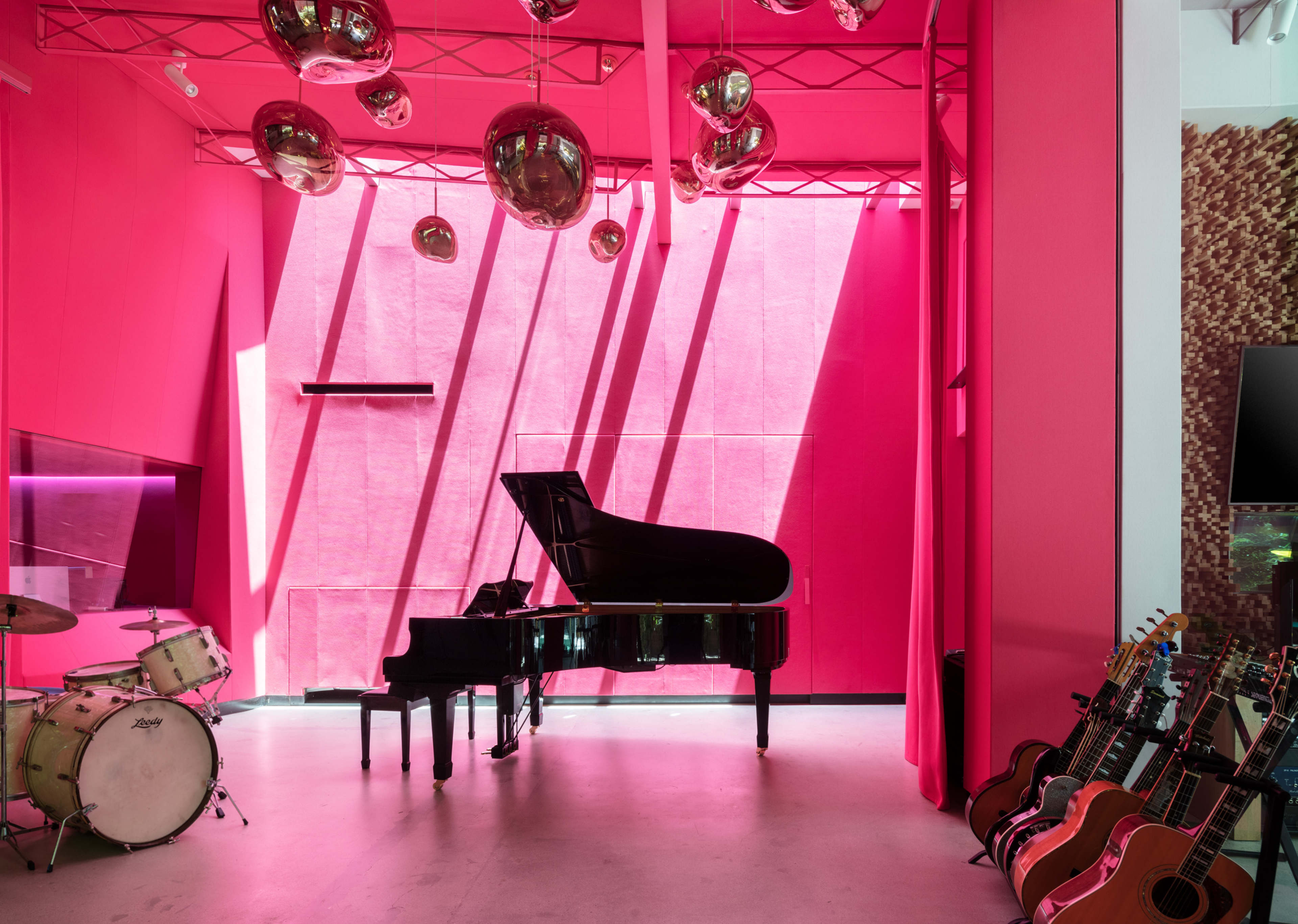 The interior features a grand piano and a drum set against a bright pink wall, with hanging metallic orbs and guitars lined up to the side.