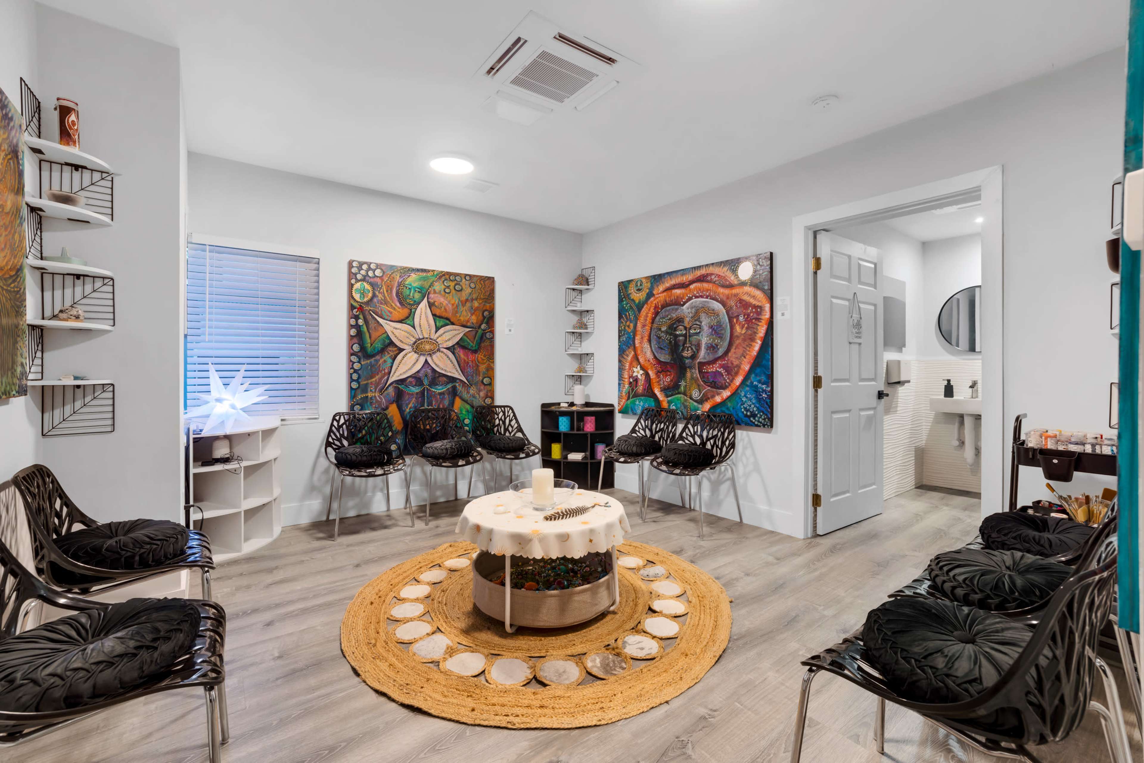 The image depicts a cozy waiting room with modern, black chairs arranged around a circular table, decorated with art on the walls and a bathroom door visible in the background.
