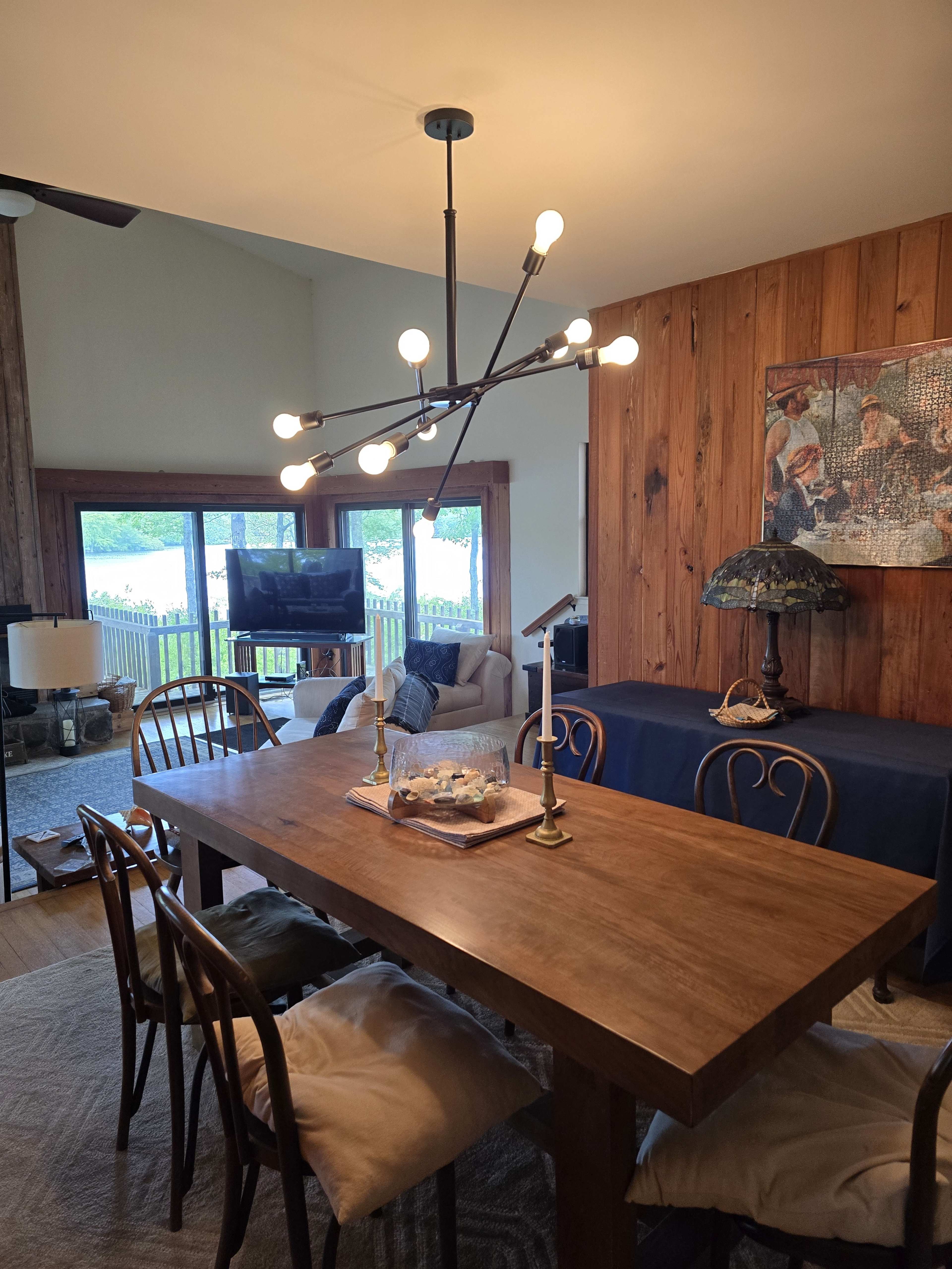 The image shows a modern dining area featuring a wooden table with chairs, a decorative chandelier, and large windows revealing a view of an outdoor space.