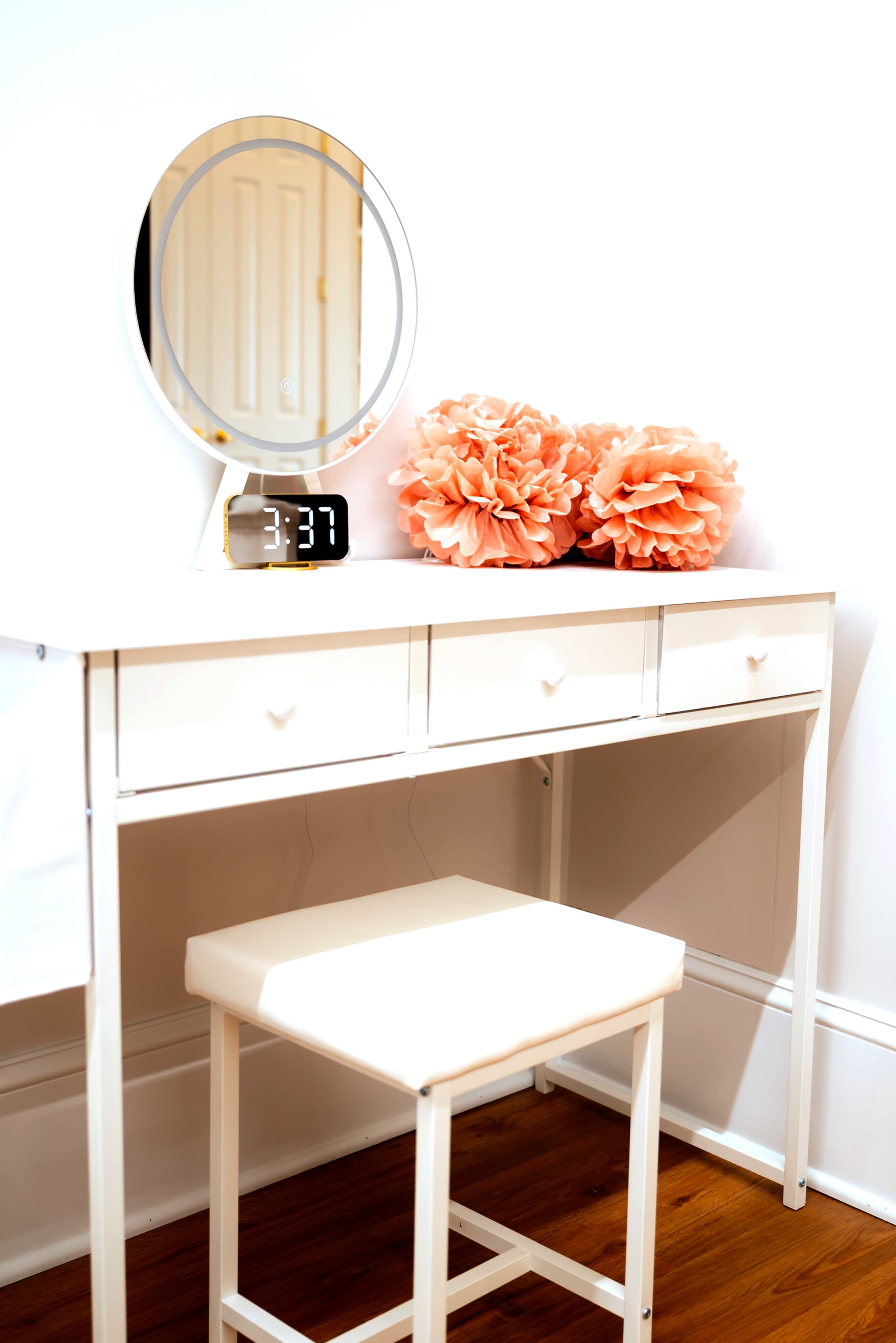 A white vanity table with three drawers, a circular mirror, an alarm clock displaying 3:37, and two large peach-colored flower decorations sits against a white wall.
