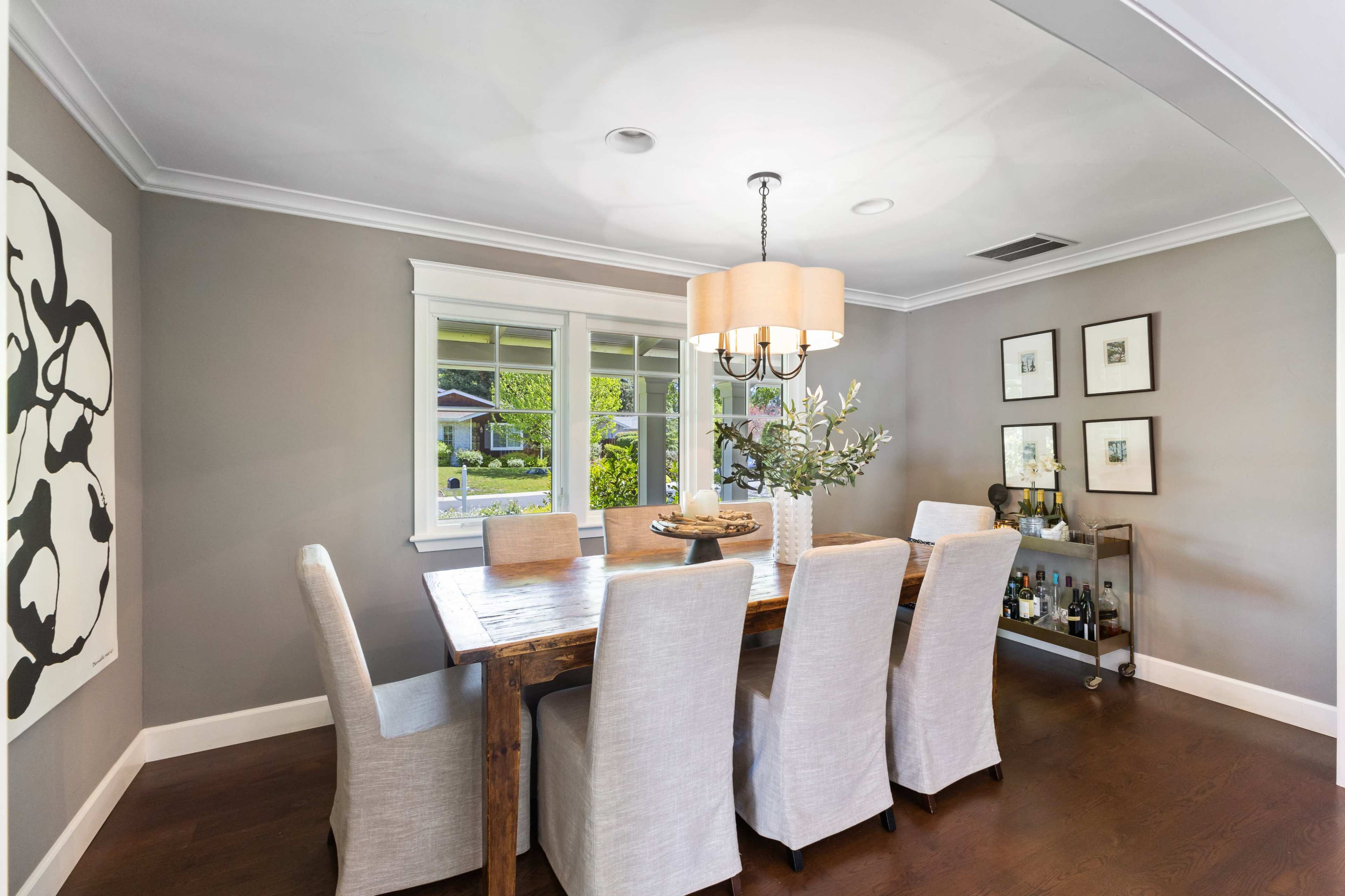 A dining room with a wooden table surrounded by beige upholstered chairs, a chandelier above, and a side cart displaying bottles and decor items.
