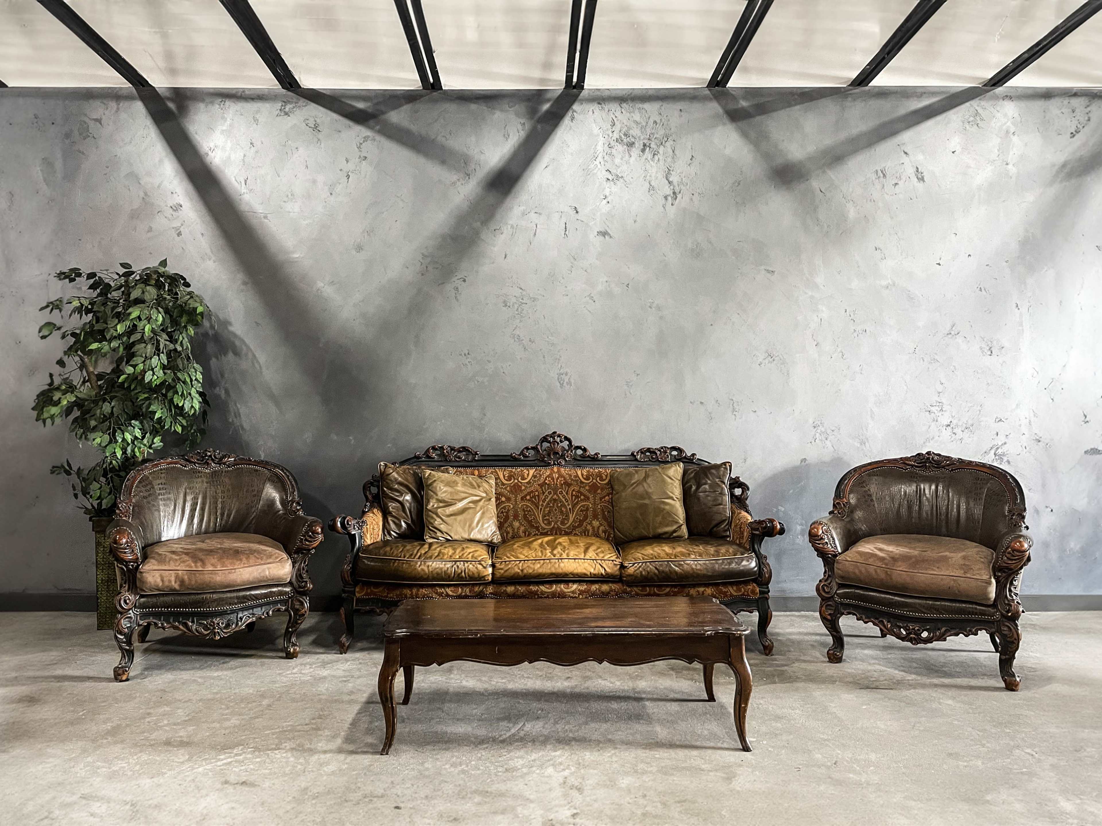 A vintage seating arrangement features a central ornate sofa flanked by two matching armchairs, set against a gray wall with a potted plant nearby.