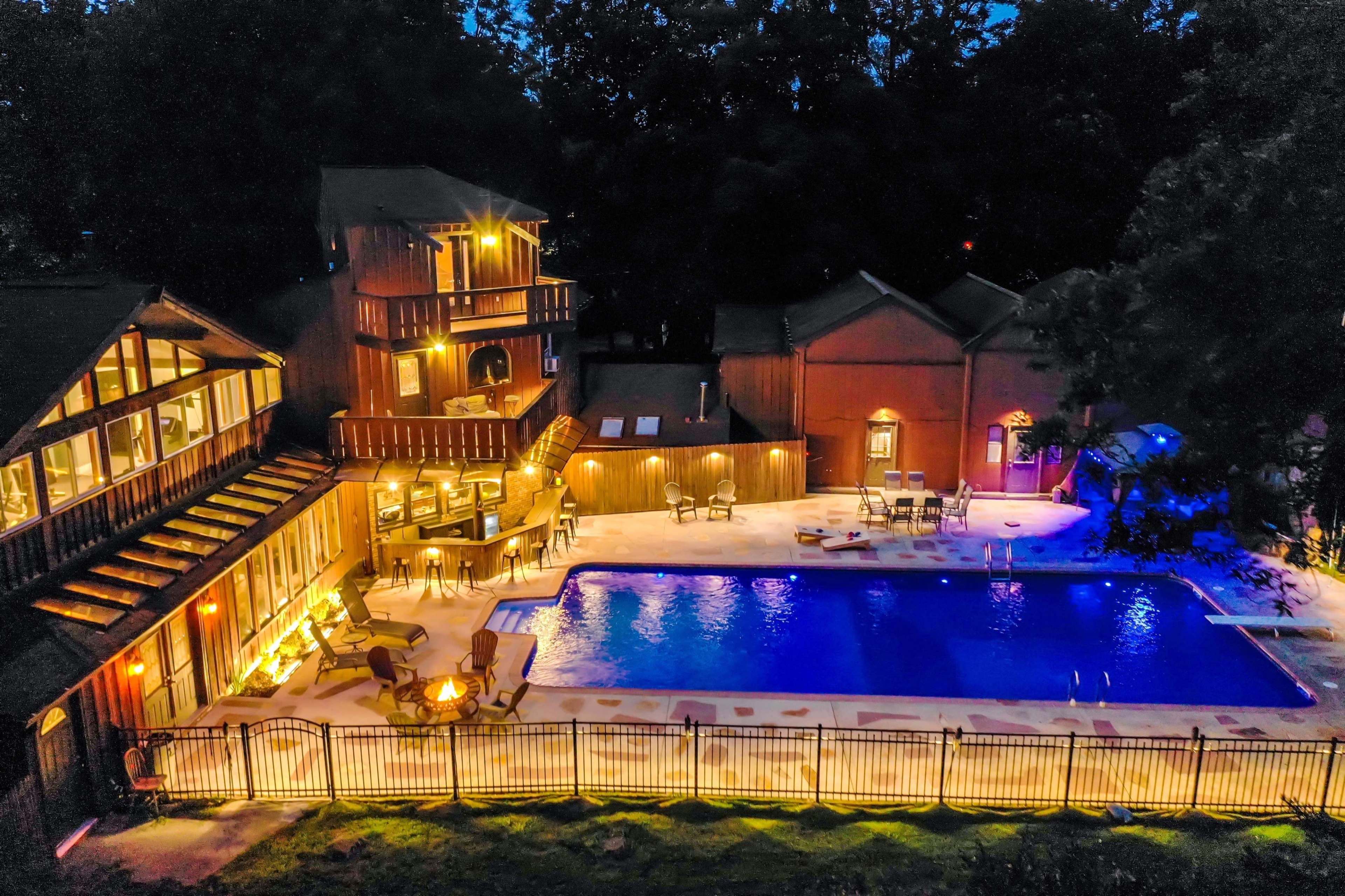 A well-lit outdoor area featuring a swimming pool surrounded by chairs and tables, with multi-storied wooden buildings in the background.