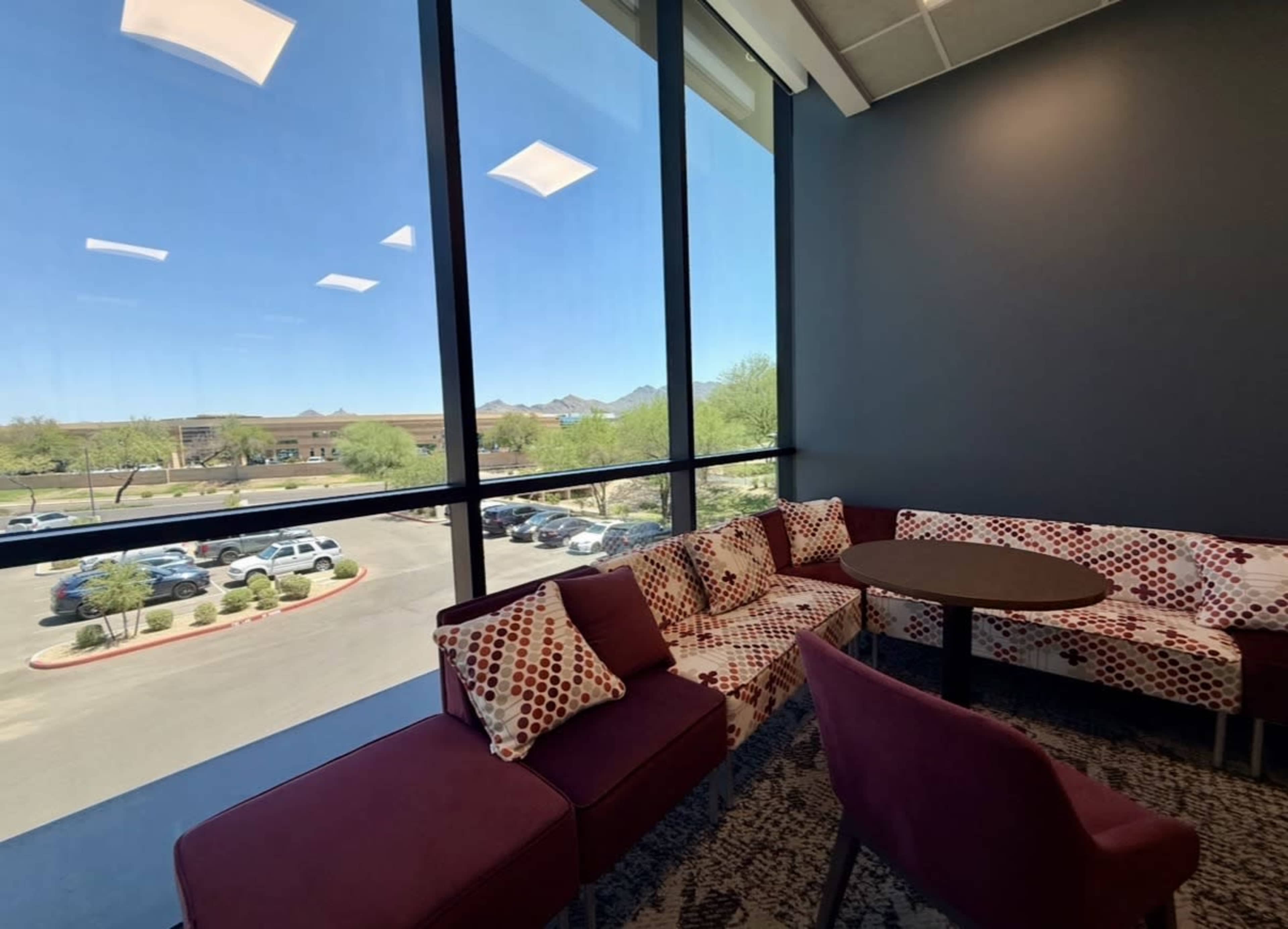 The image shows a lounge area with a circular table and patterned seating, overlooking a parking lot and distant mountains through large windows.