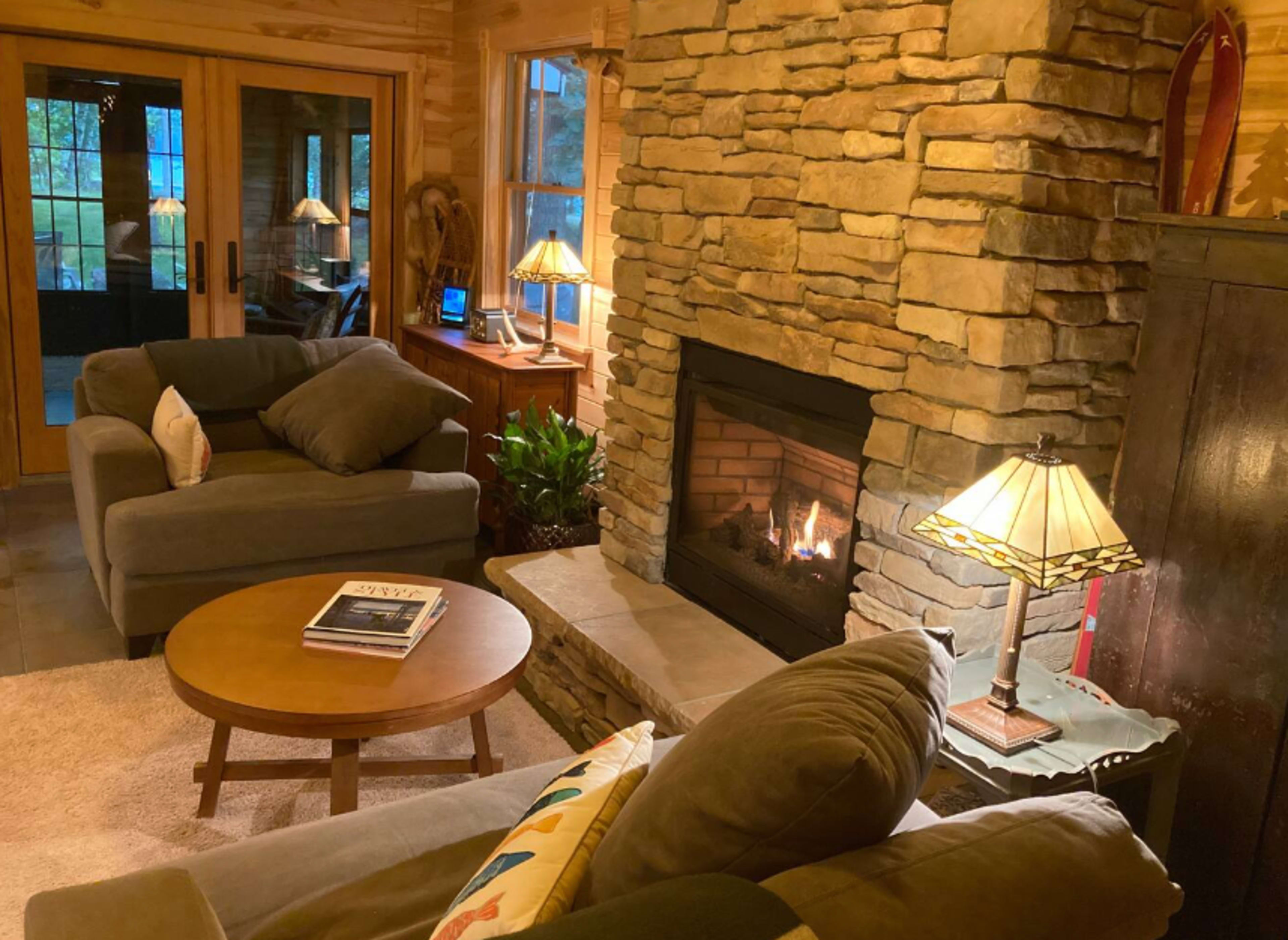 A cozy living room features a stone fireplace, two gray couches, a wooden coffee table, and warm lighting from lamps.