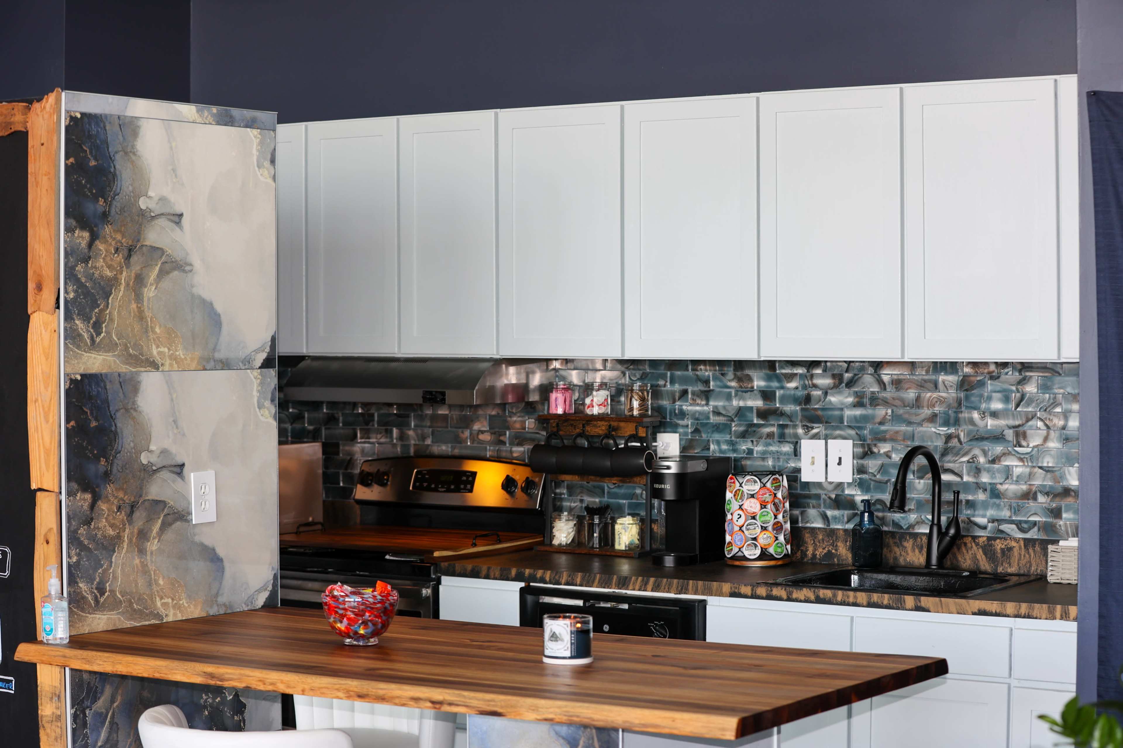 The image shows a modern kitchen featuring white cabinets, a dark countertop, and a backsplash made of blue and gray tiles.