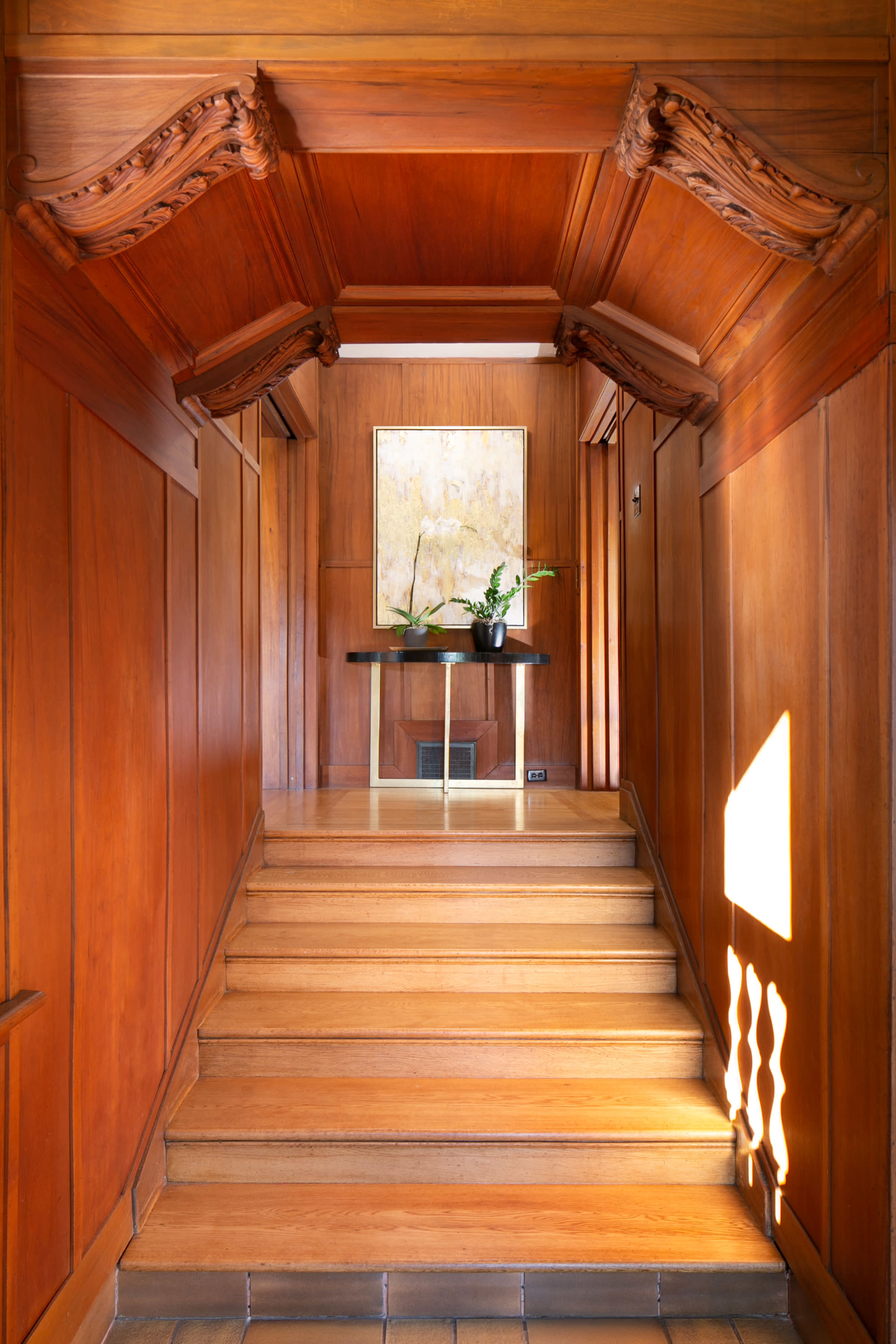A wooden hallway with a staircase leading up to a landing, featuring a small table and a wall-mounted mirror.