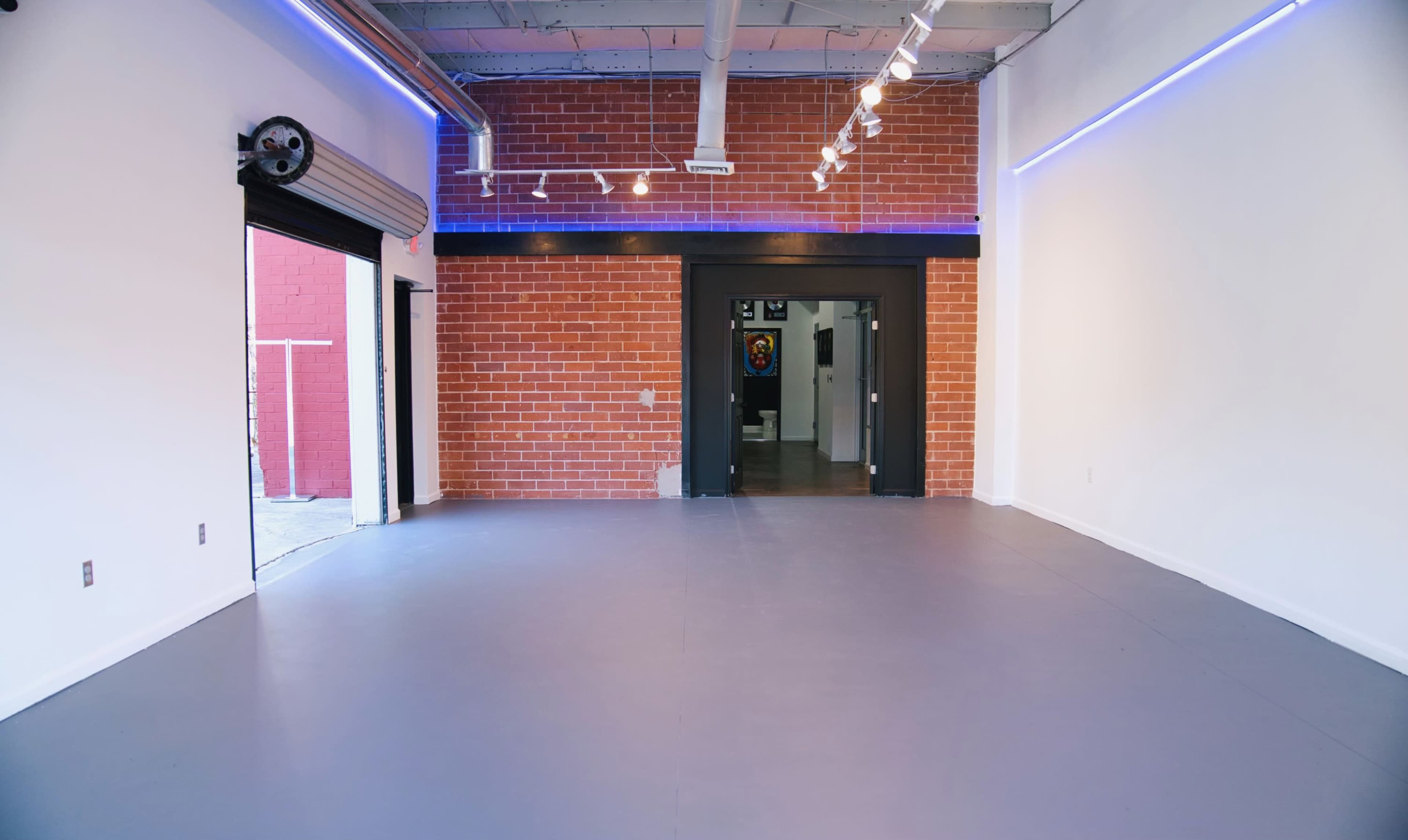 An empty interior space with a polished gray floor, exposed brick walls, and a large black door at the back leading to another room.