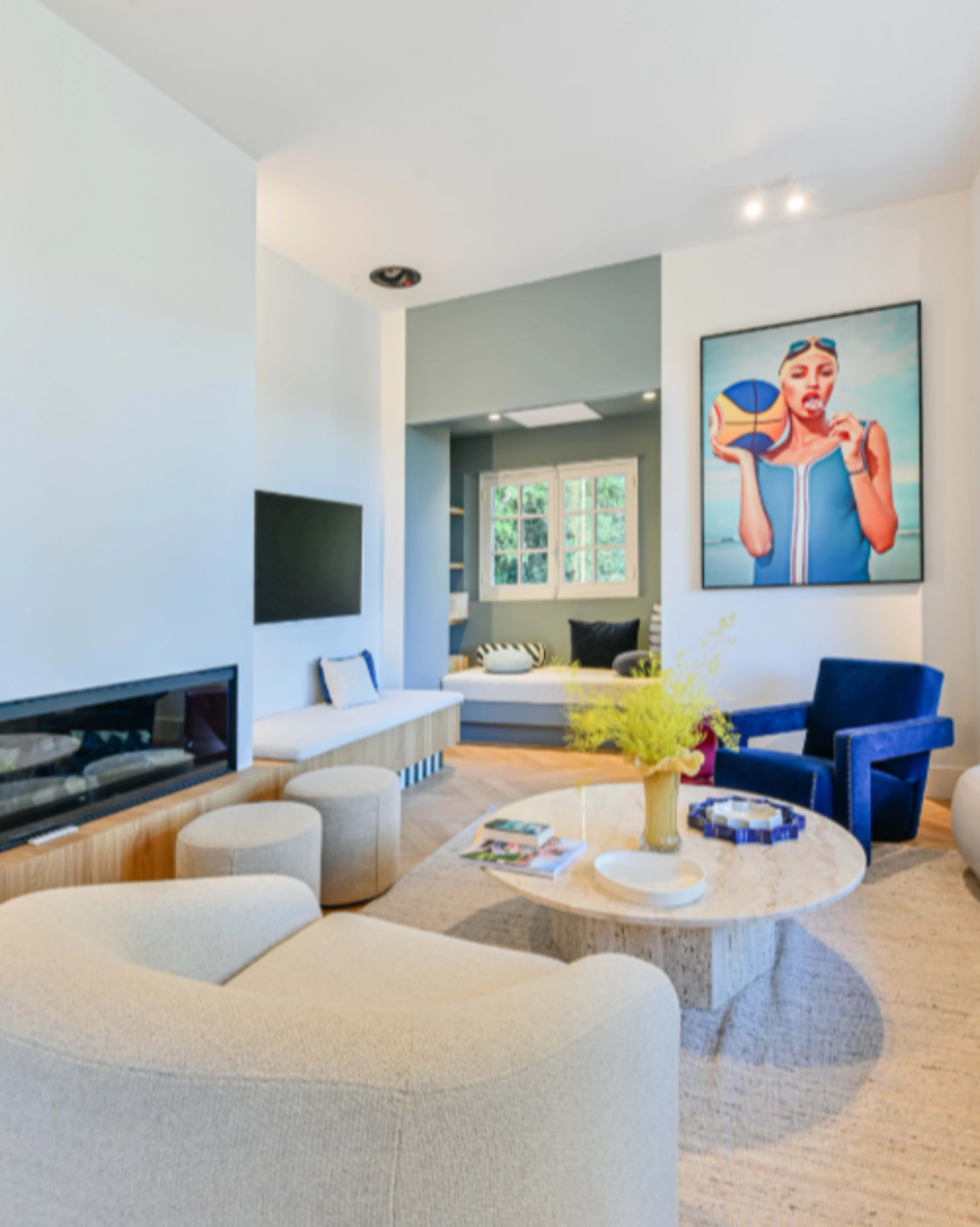 The image shows a modern living room featuring a fireplace, a large wall art piece, comfortable seating, and natural light coming through windows.