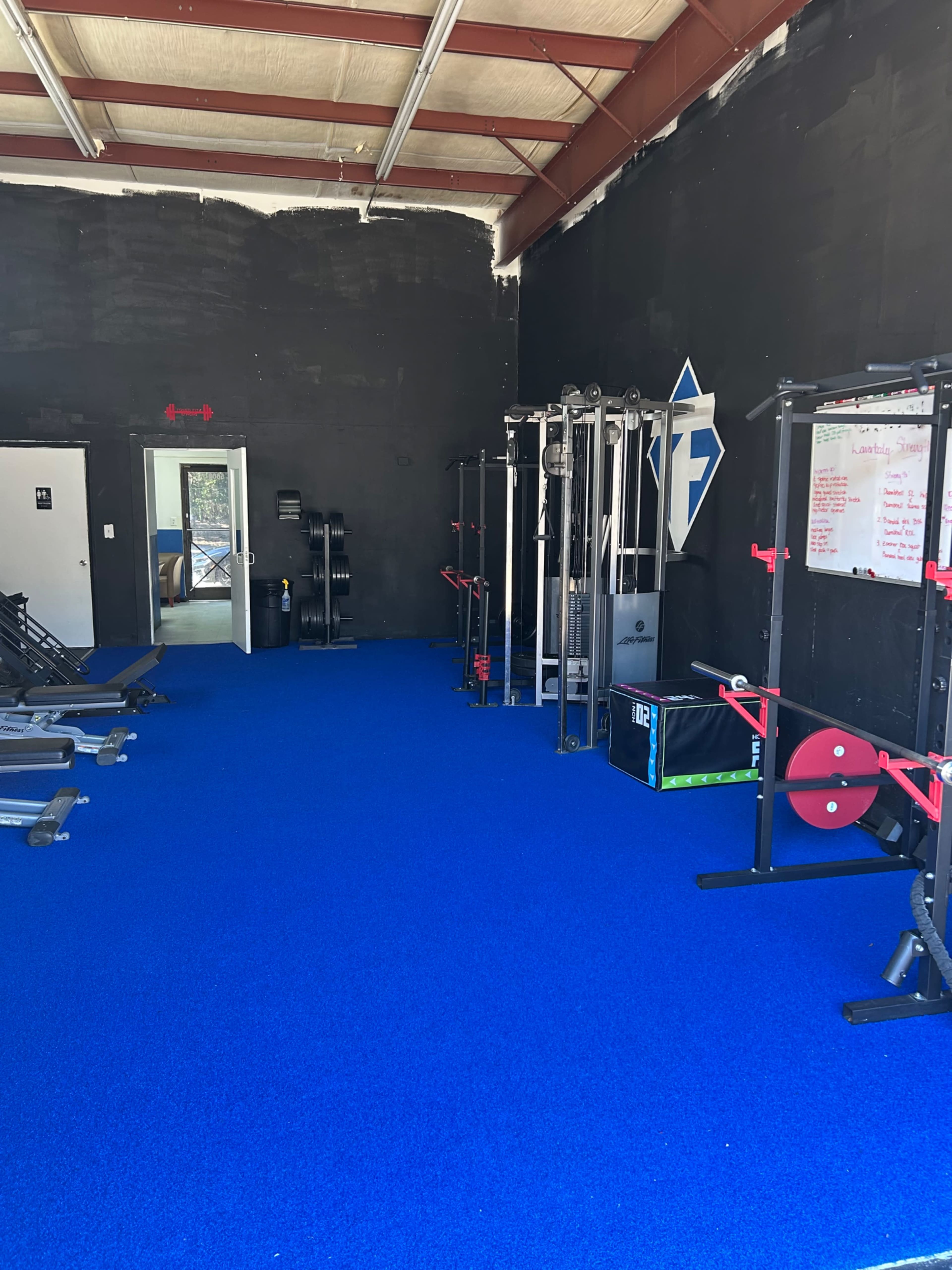 The image shows a fitness studio with blue flooring, exercise equipment lined against the walls, and a whiteboard with notes in the background.