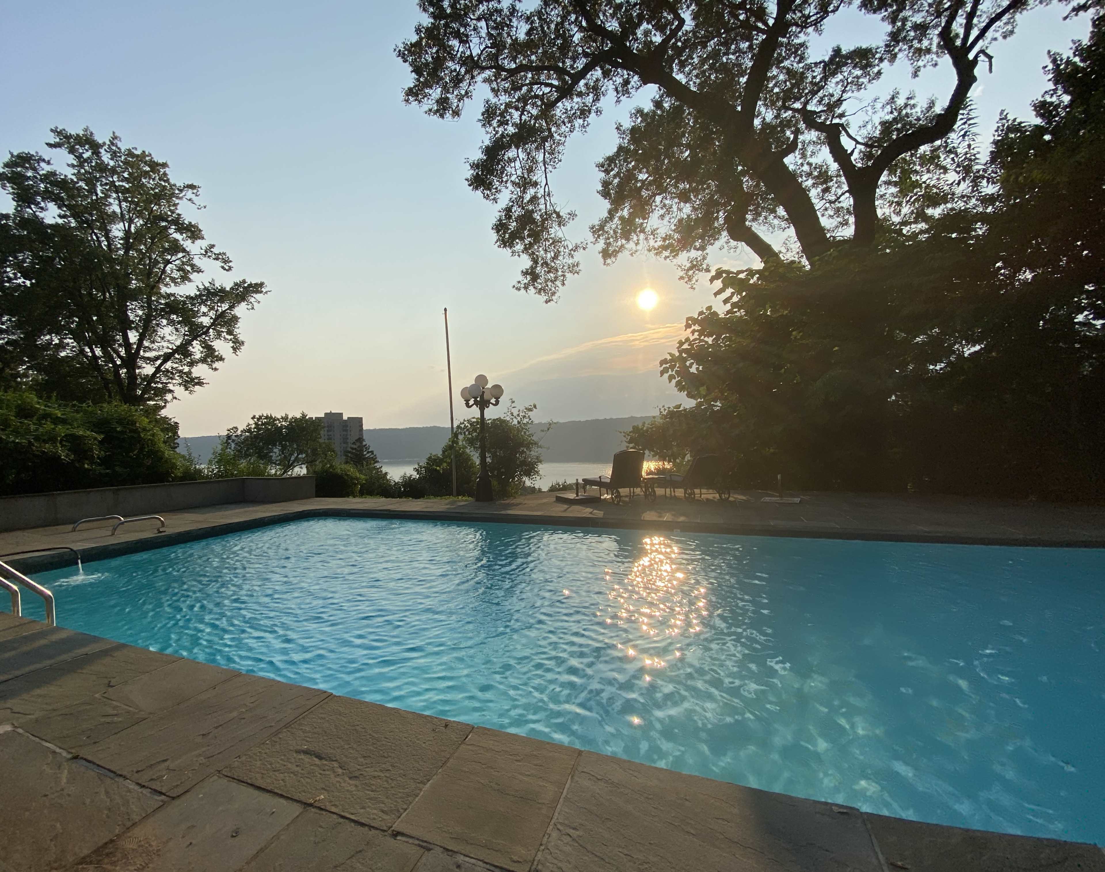 The image shows a swimming pool surrounded by trees, with the sun setting in the background over a body of water.