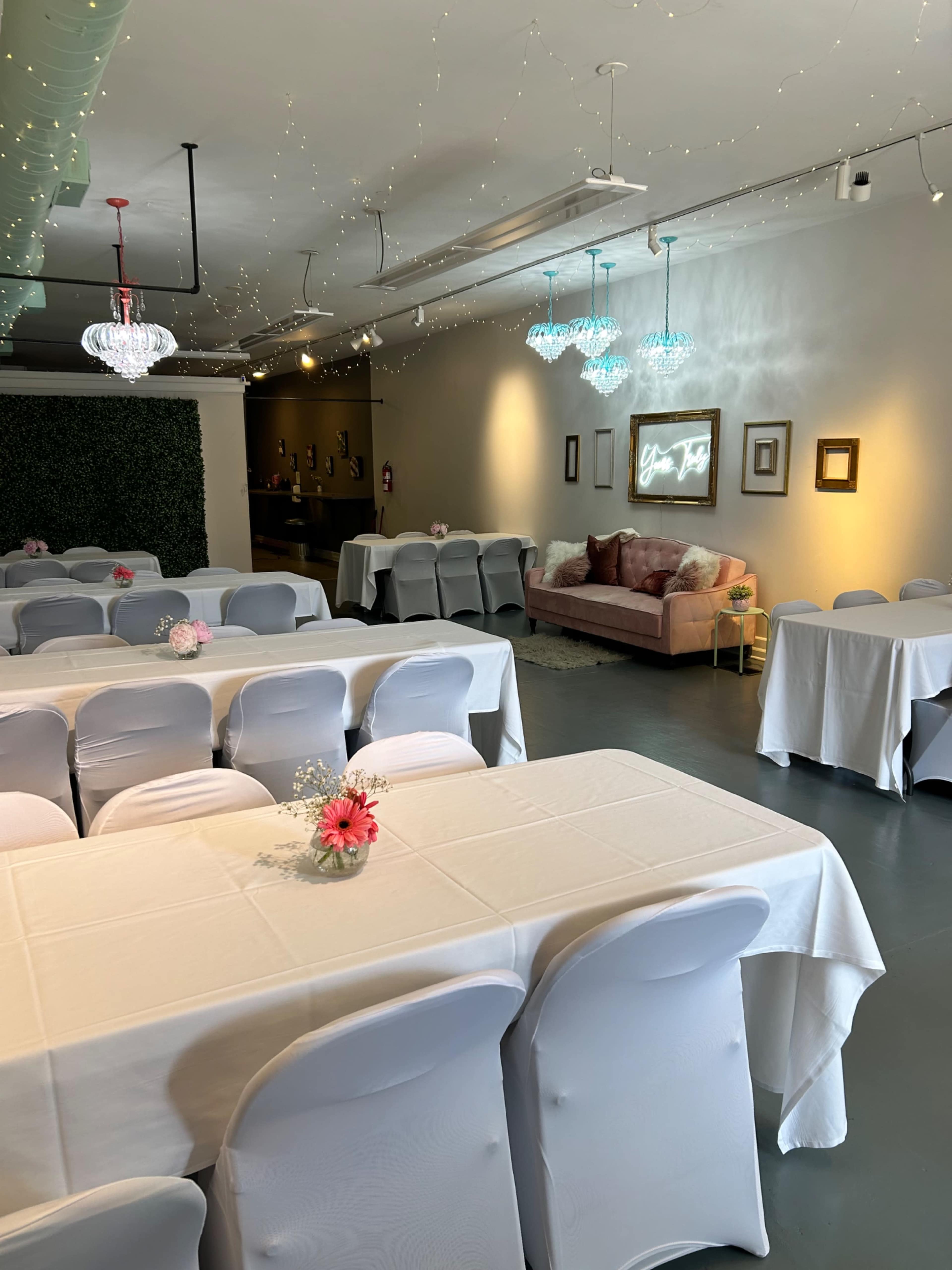 A spacious event room with neatly arranged tables covered in white tablecloths, and a cozy seating area adorned with decorative lights and a floral centerpiece on each table.