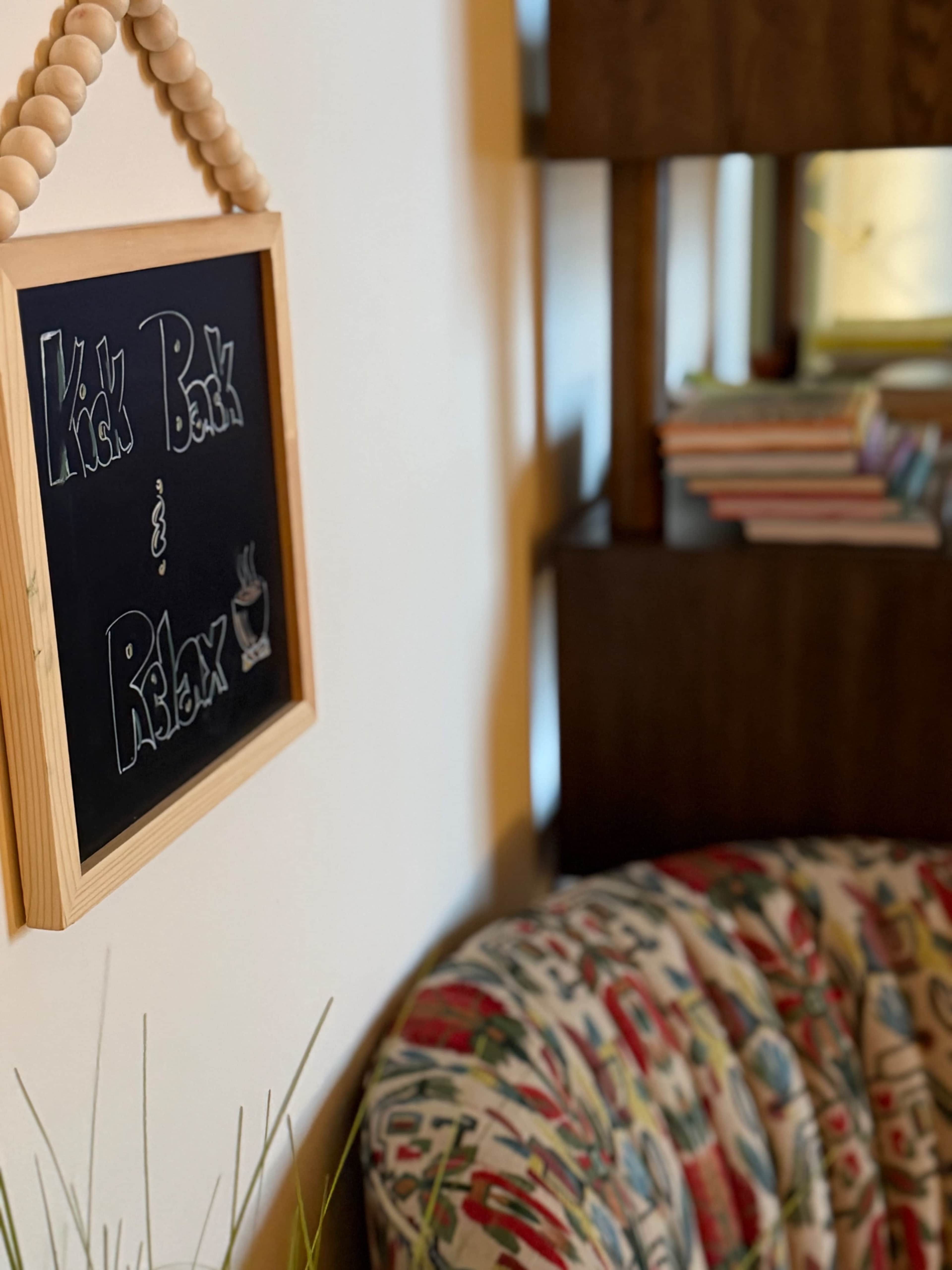 A wooden sign that reads "Kick Back & Relax" hangs on a wall next to a patterned, cushioned chair and a shelf with books.
