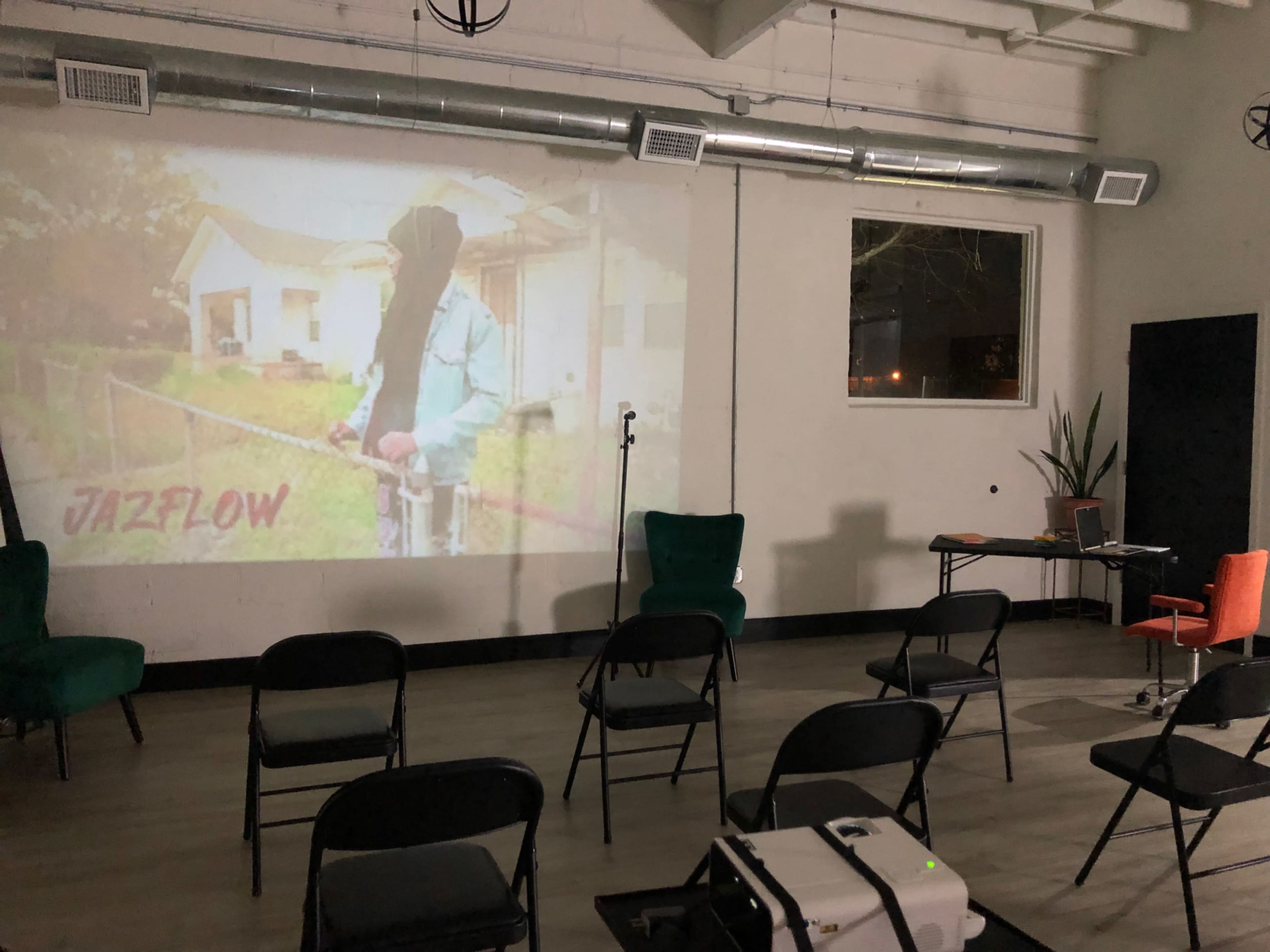 The scene shows a dimly lit room set up for a presentation, featuring chairs arranged in rows, a projector displaying an image of a person, and a small stage with a microphone.