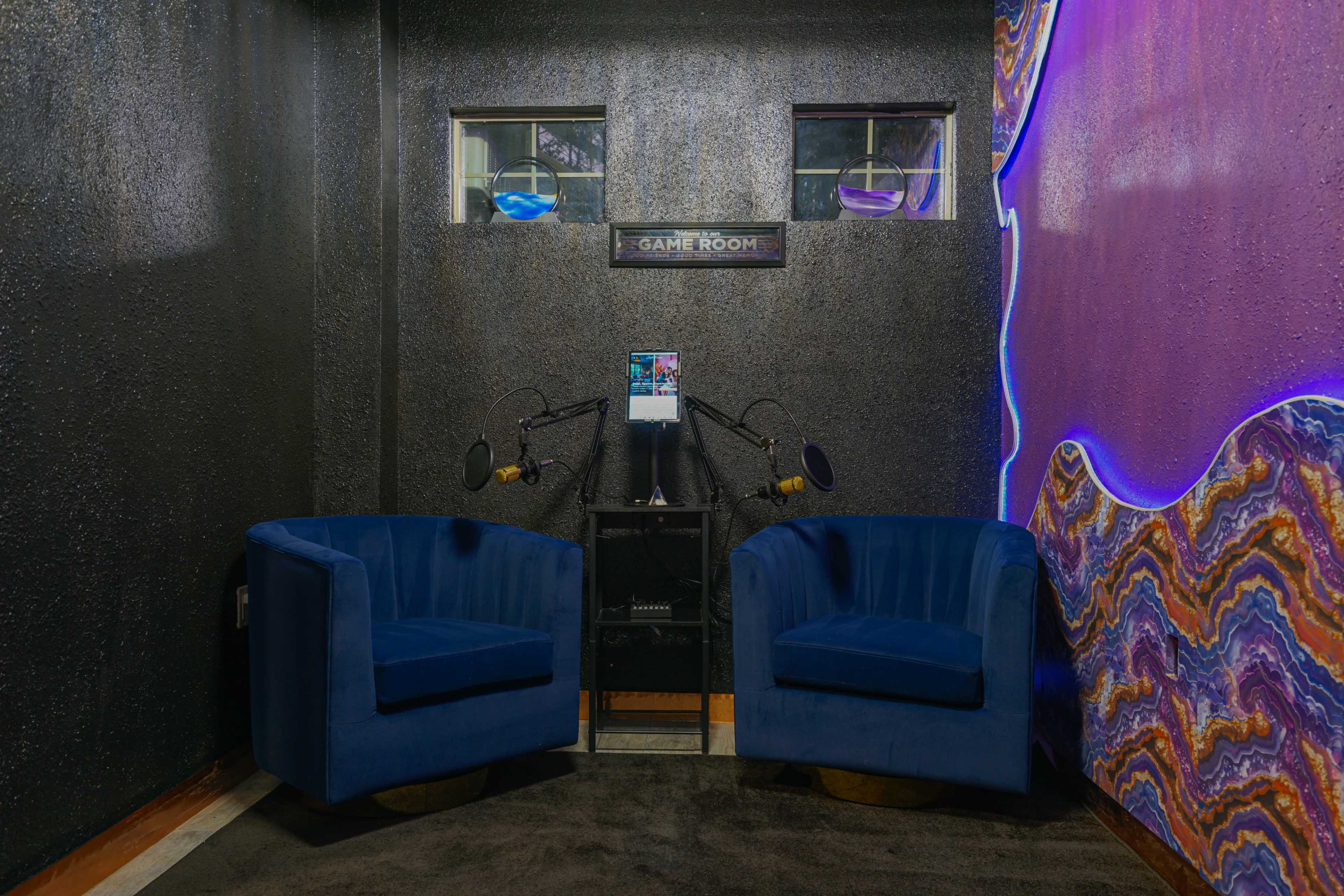 The image shows a small game room featuring two blue velvet chairs, a black shelf with a tablet, and colorful lighting on the walls.