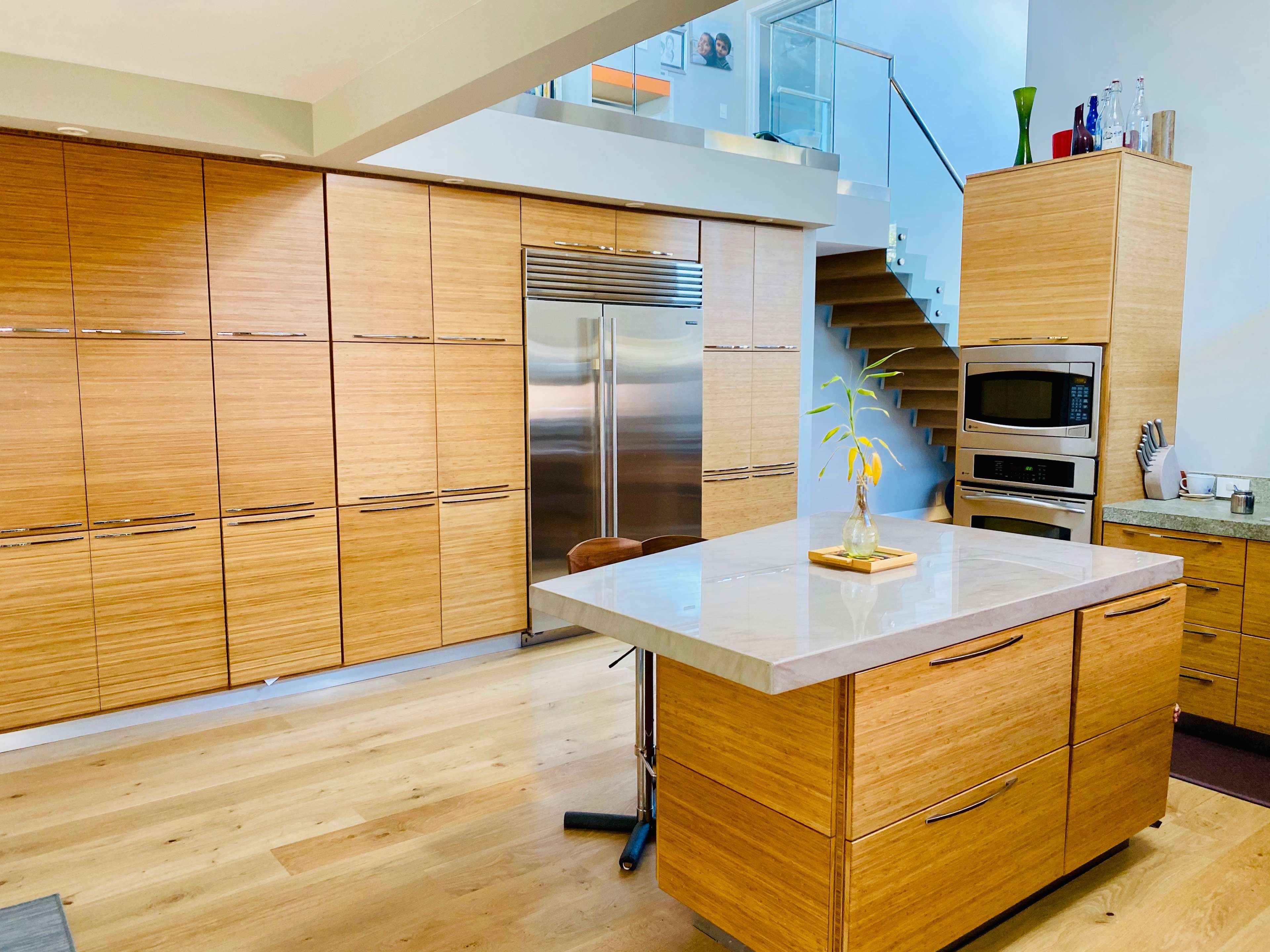 A modern kitchen with wooden cabinetry, a stainless steel refrigerator, and a central island topped with a light-colored stone surface.