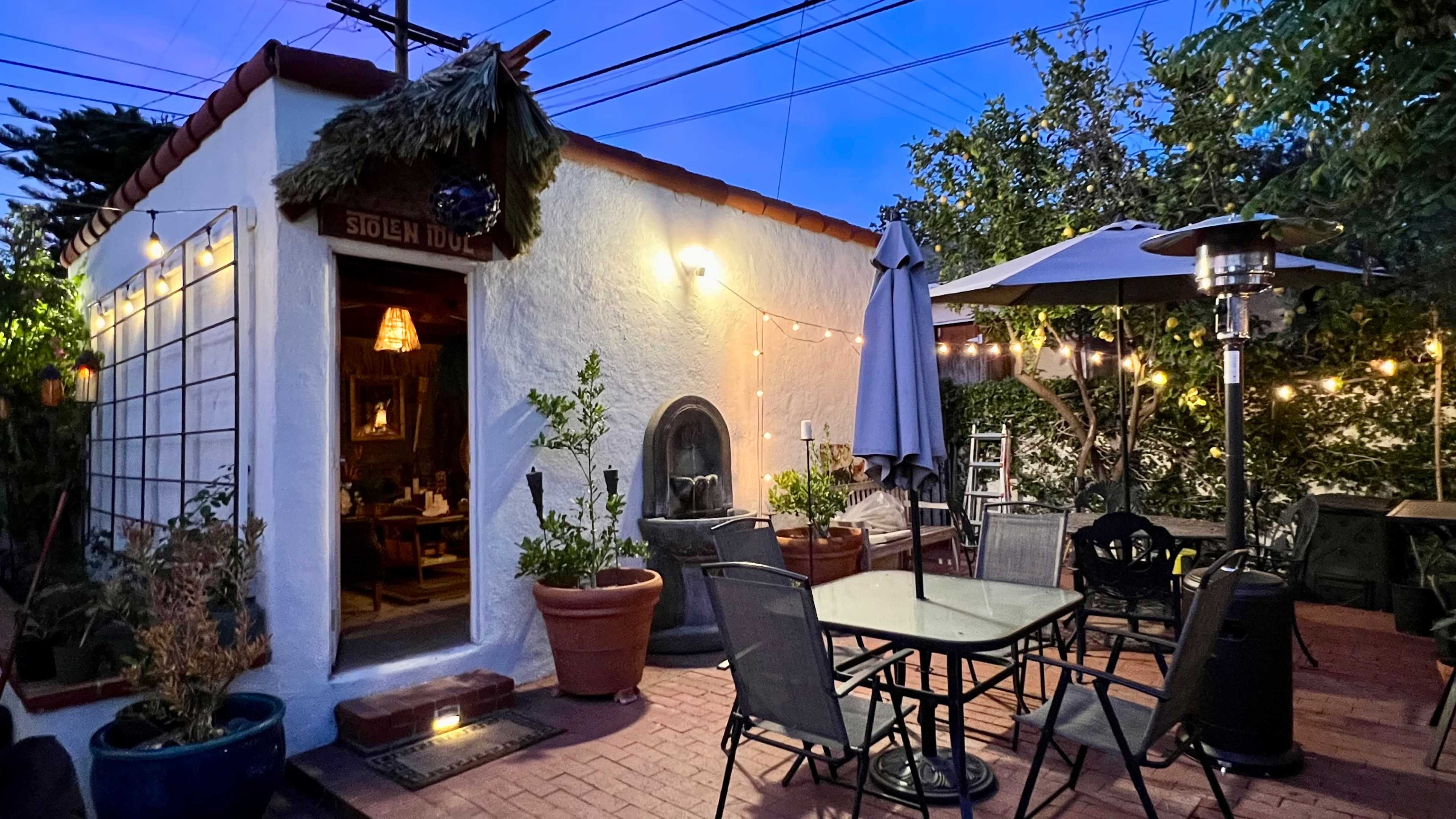 The outdoor patio features a table and chairs surrounded by potted plants, with a small building in the background illuminated by hanging lights.