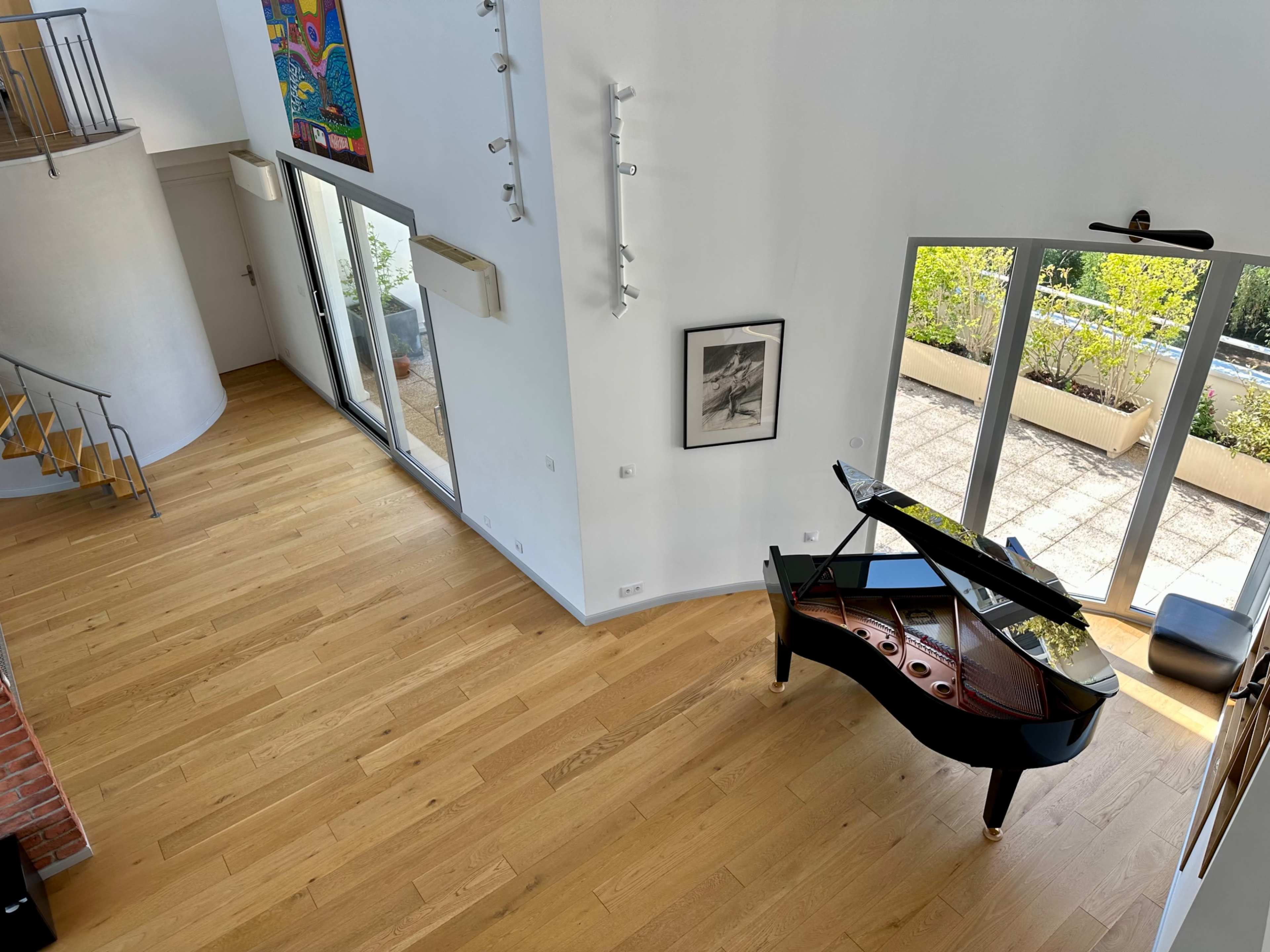 The image shows a spacious interior with a grand piano, large windows, and wood flooring, overlooking an outdoor patio area.