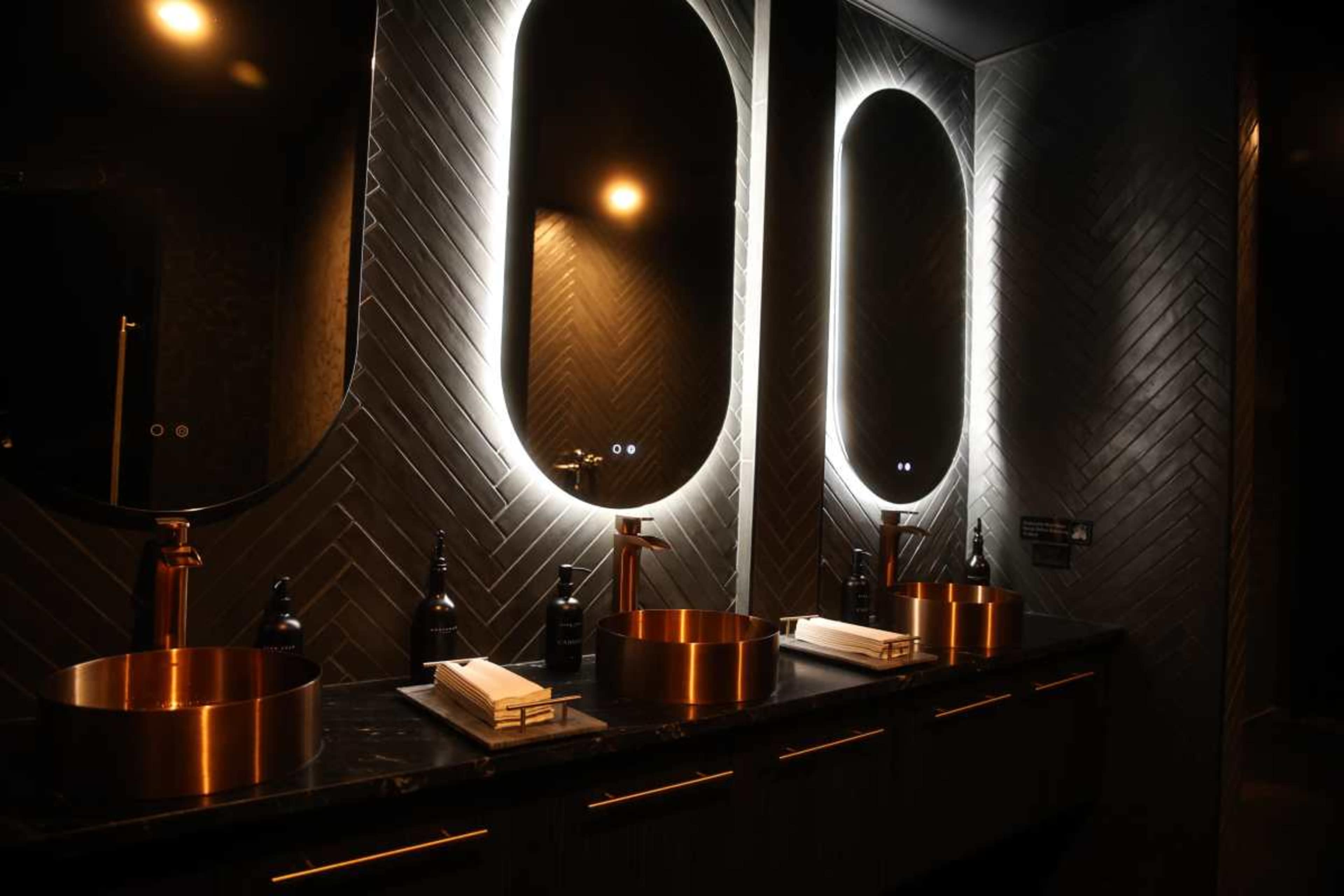 The image shows a modern bathroom with three circular mirrors illuminated by soft lighting above three copper sinks on a dark countertop.