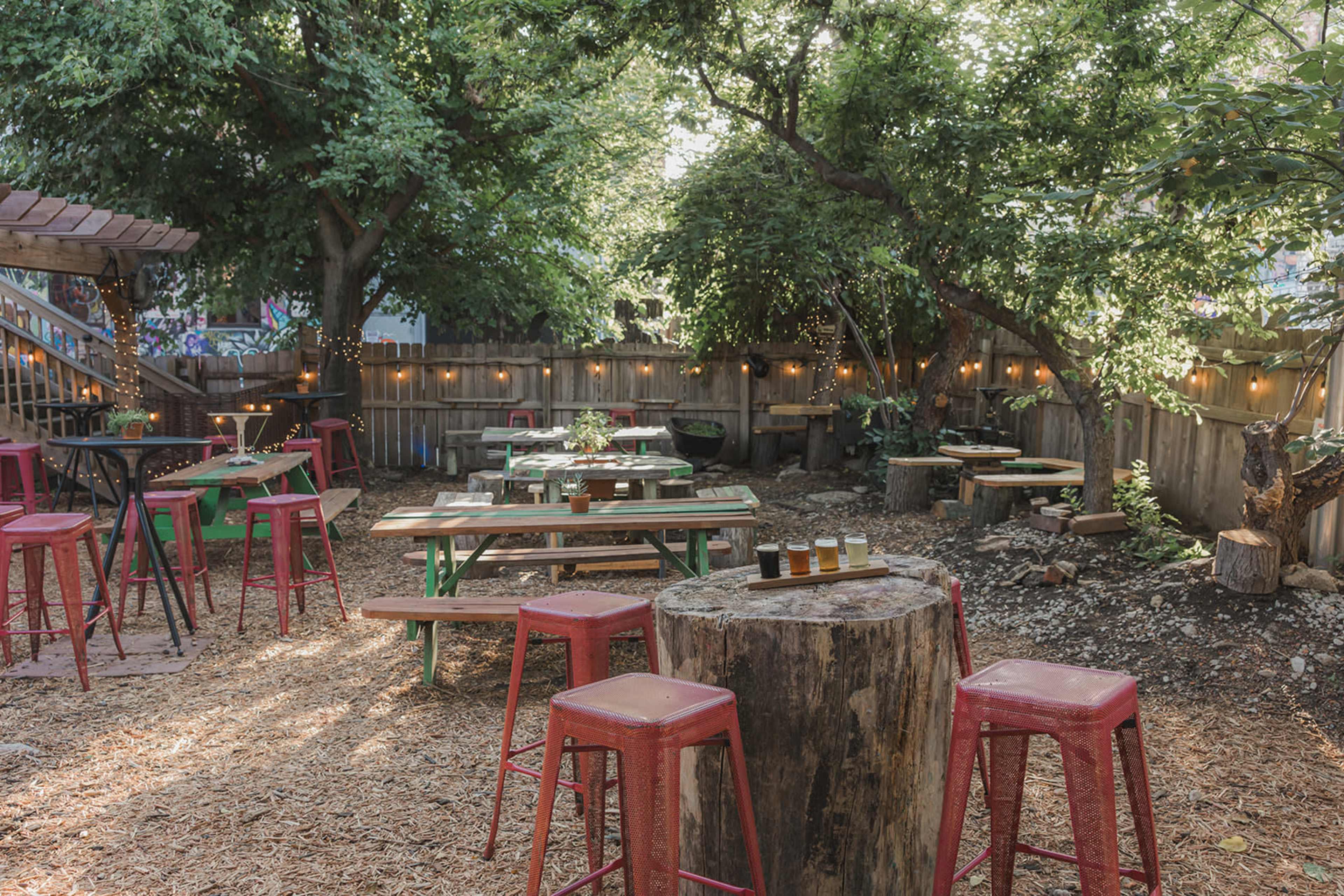The image shows an outdoor seating area with wooden tables and red metal stools surrounded by trees and string lights in a gravel setting.