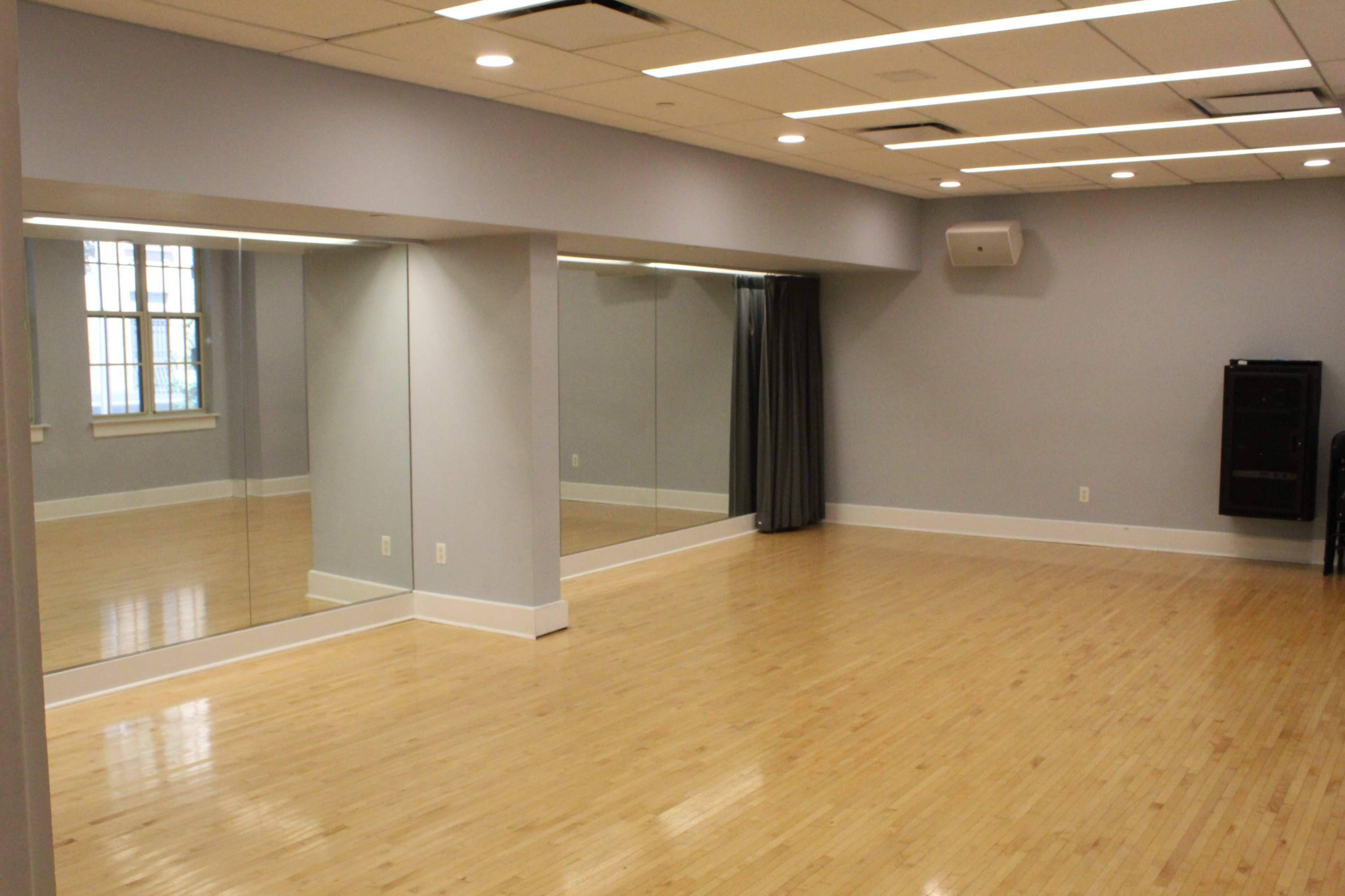 The image shows a spacious, empty room with wooden flooring, large mirrors on one wall, and a gray color scheme adorned with soft lighting.