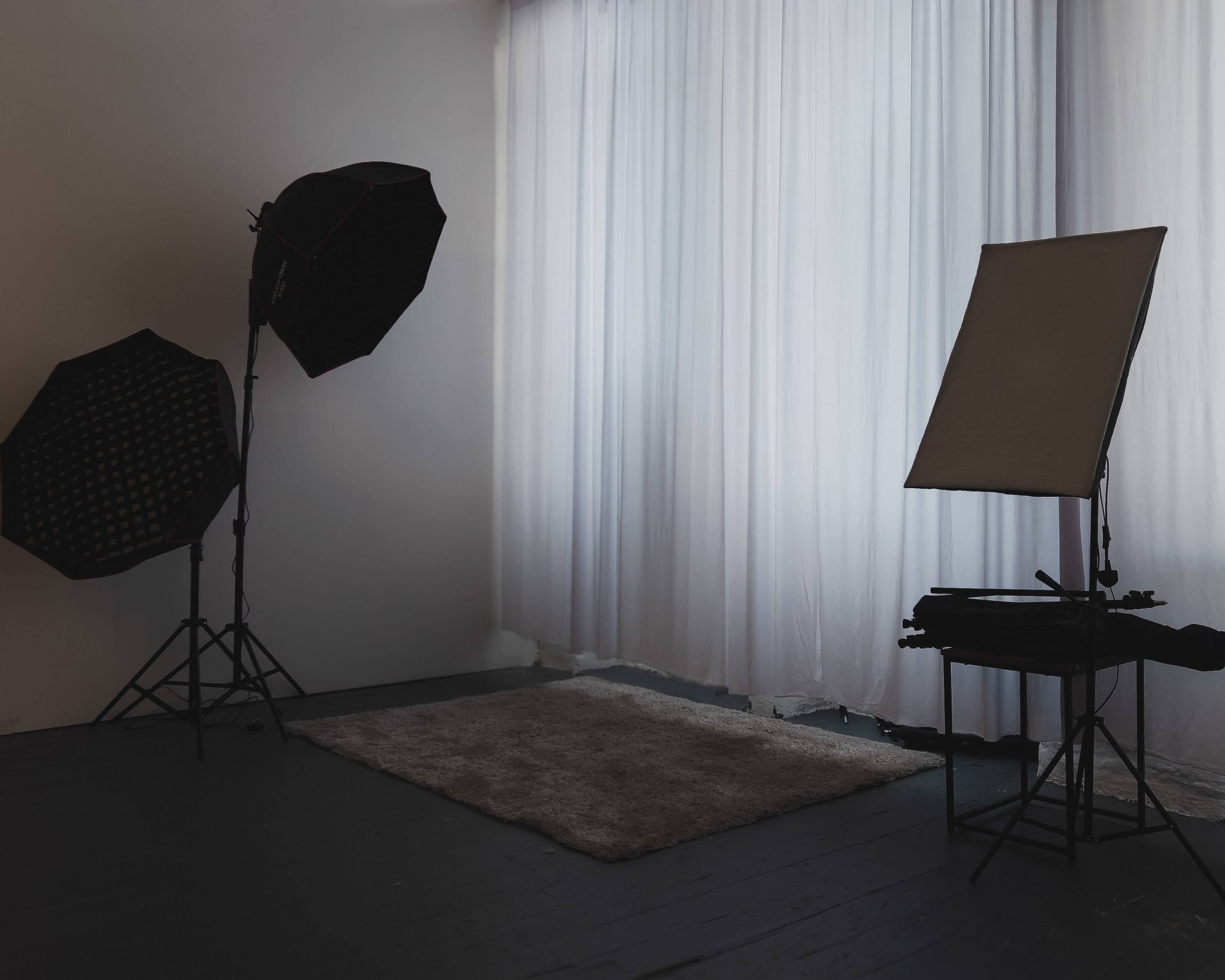 The image shows a photography studio with soft lighting, a rug on the floor, and various lighting equipment positioned near sheer curtains.