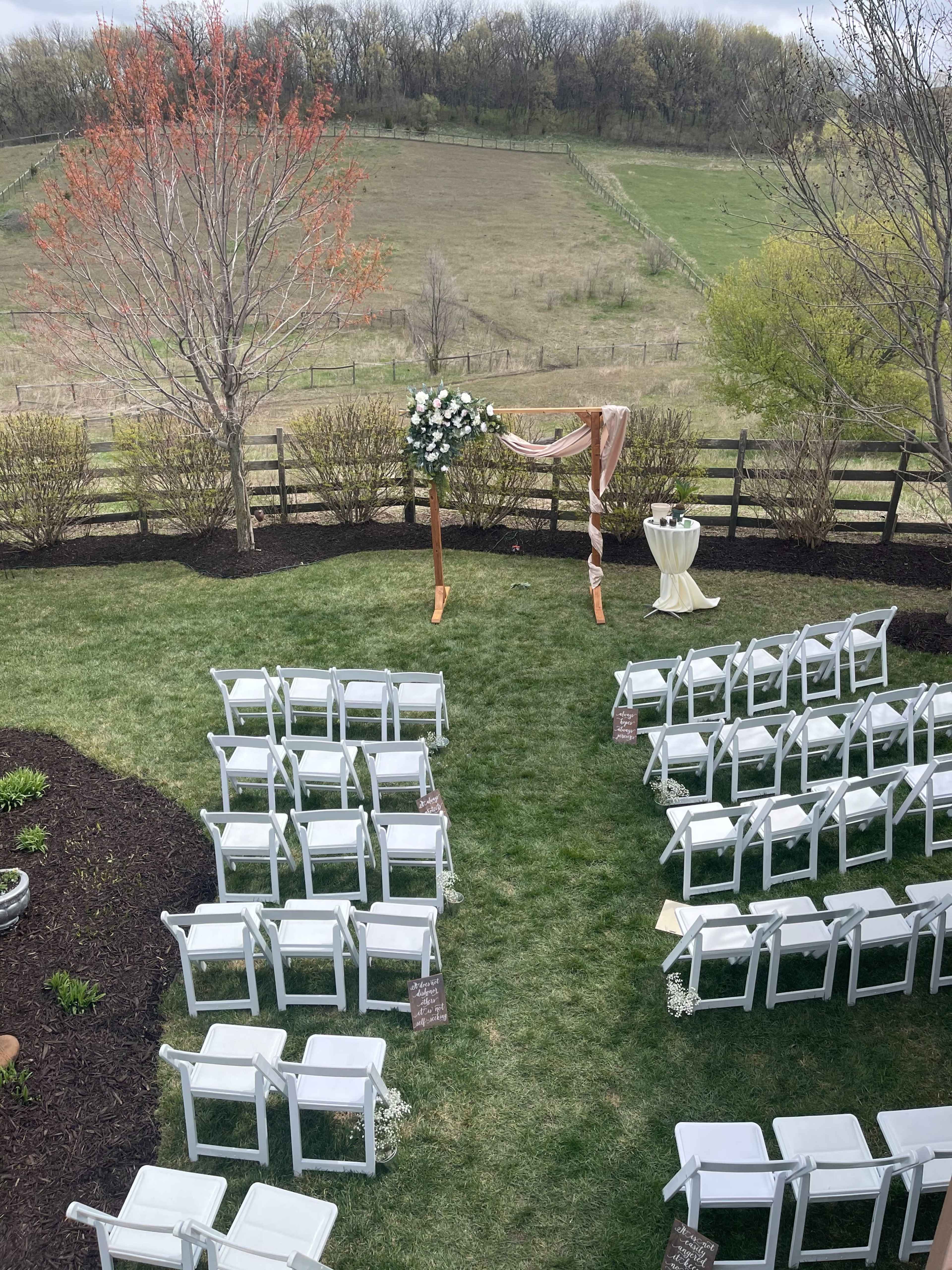 An outdoor wedding setup features white chairs arranged in rows facing an arch adorned with flowers, set against a backdrop of rolling hills and trees.