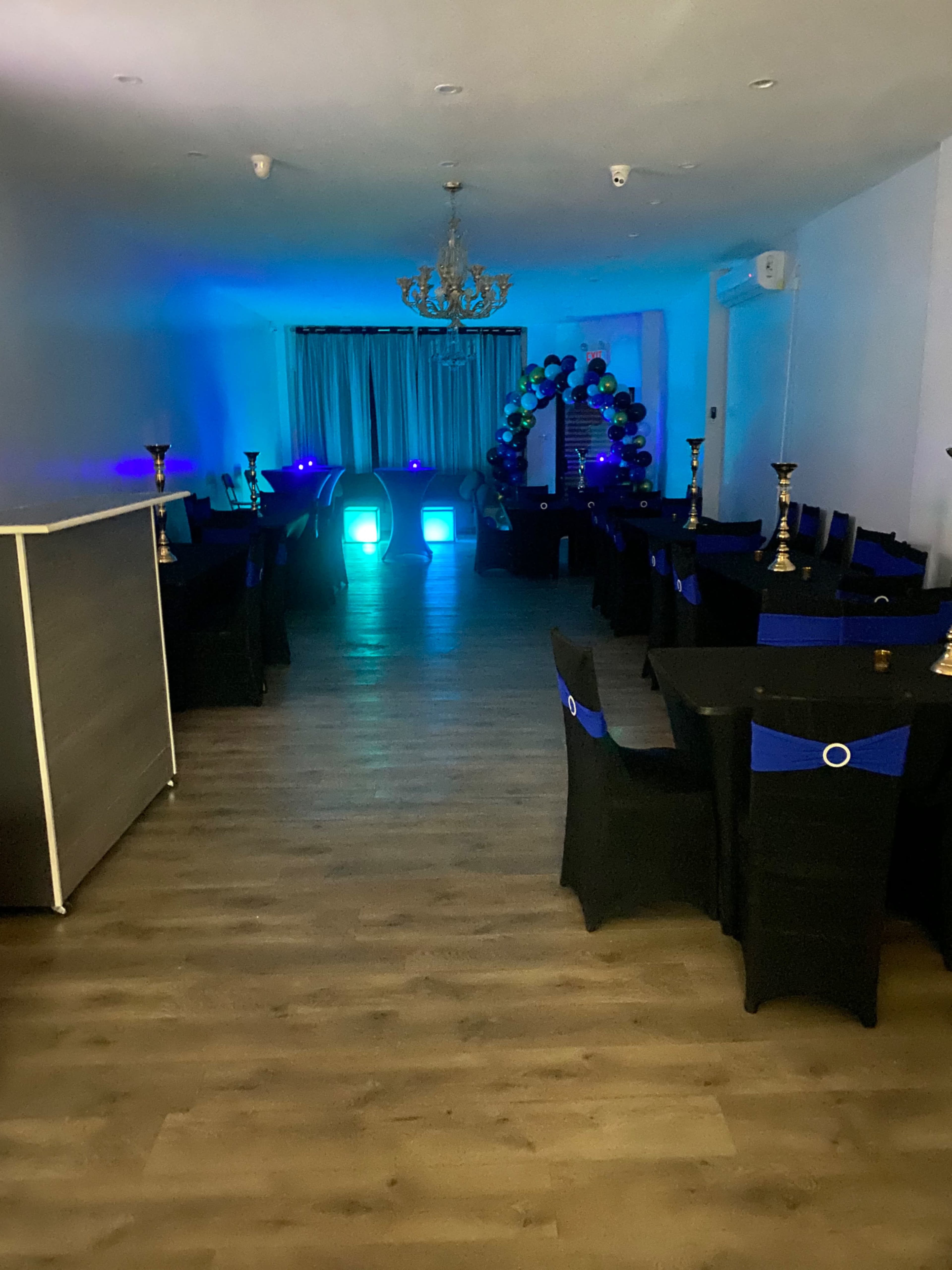 A dimly lit event space features black tablecloths with blue accents, illuminated by colored lights and decorated with a balloon arch at one end.