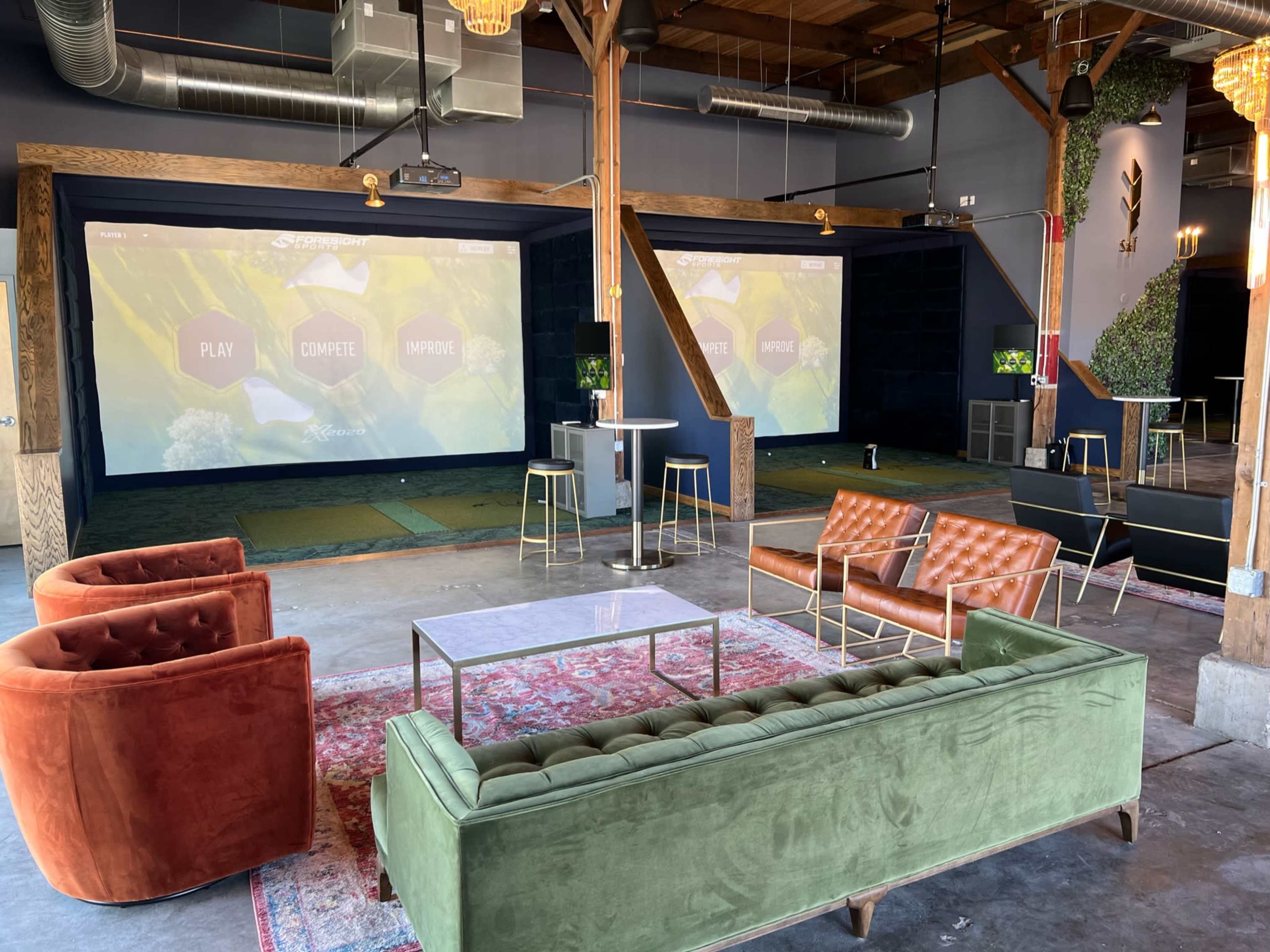 A spacious indoor lounge area featuring a green sofa, brown chairs, a glass table, and two screens displaying interactive content.