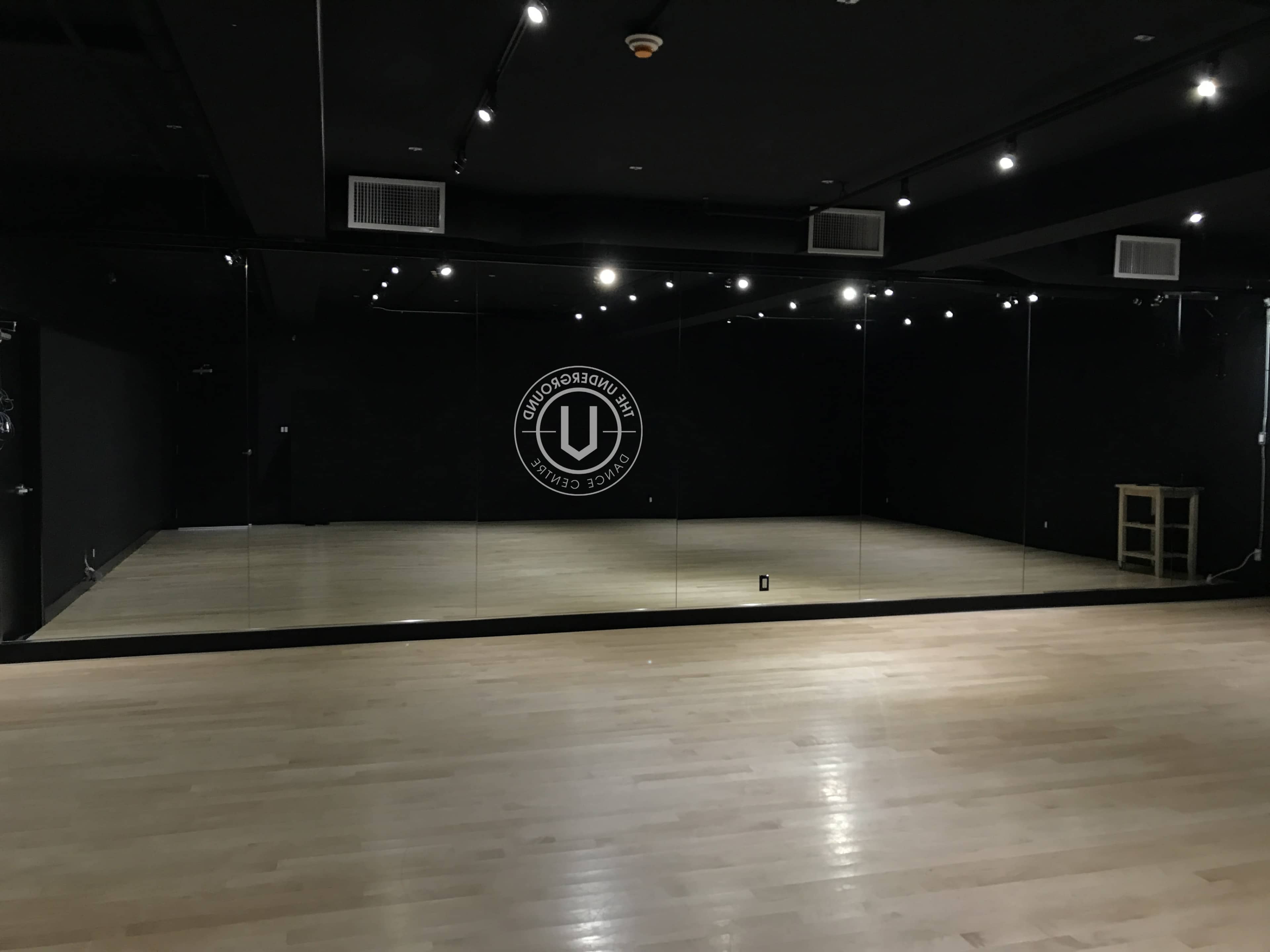 A spacious, dimly lit dance studio features polished wooden floors and a large mirrored wall with a circular logo.