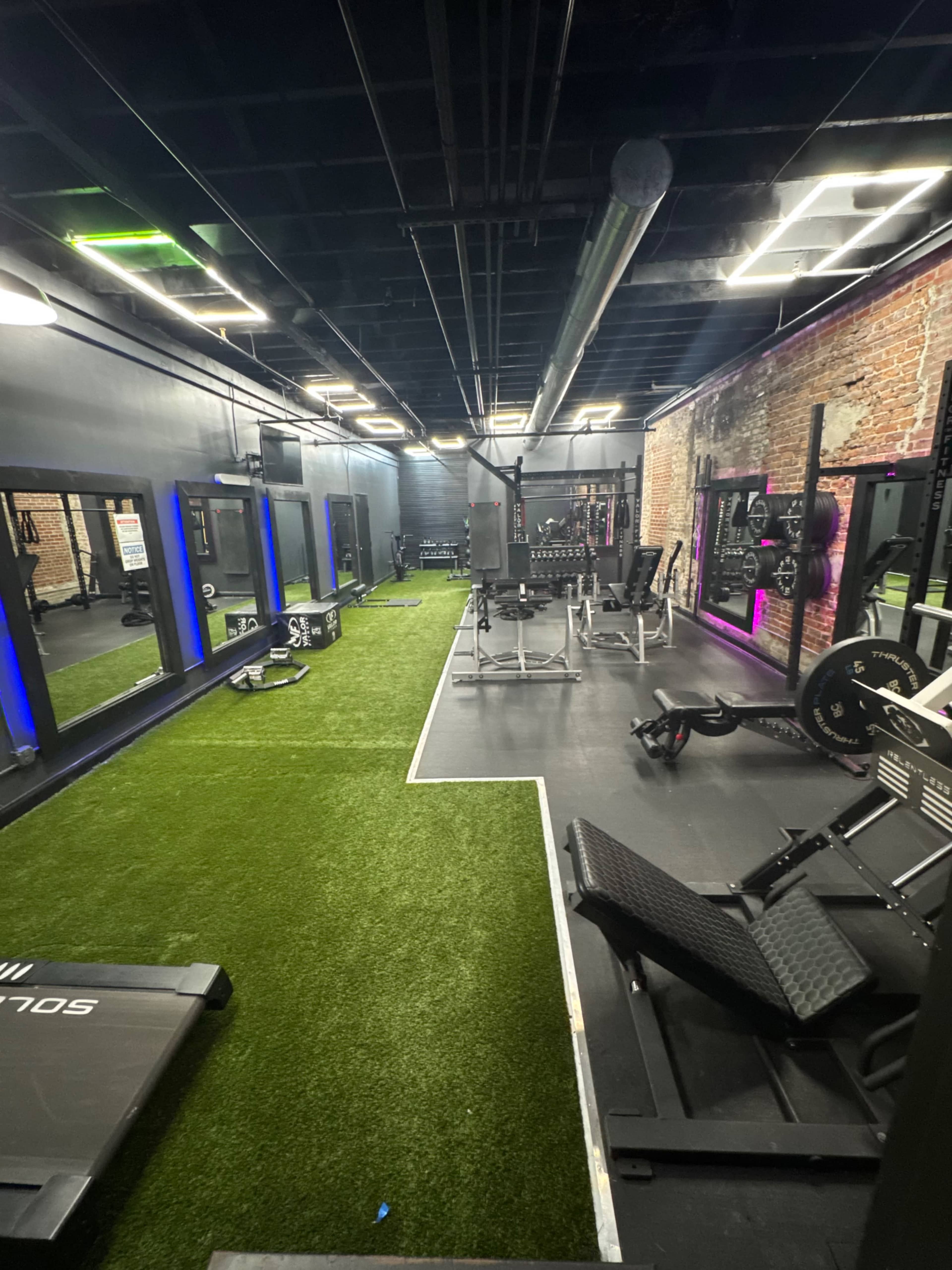 The image shows a gym interior featuring various exercise equipment, turf flooring, mirrored walls, and overhead lighting.