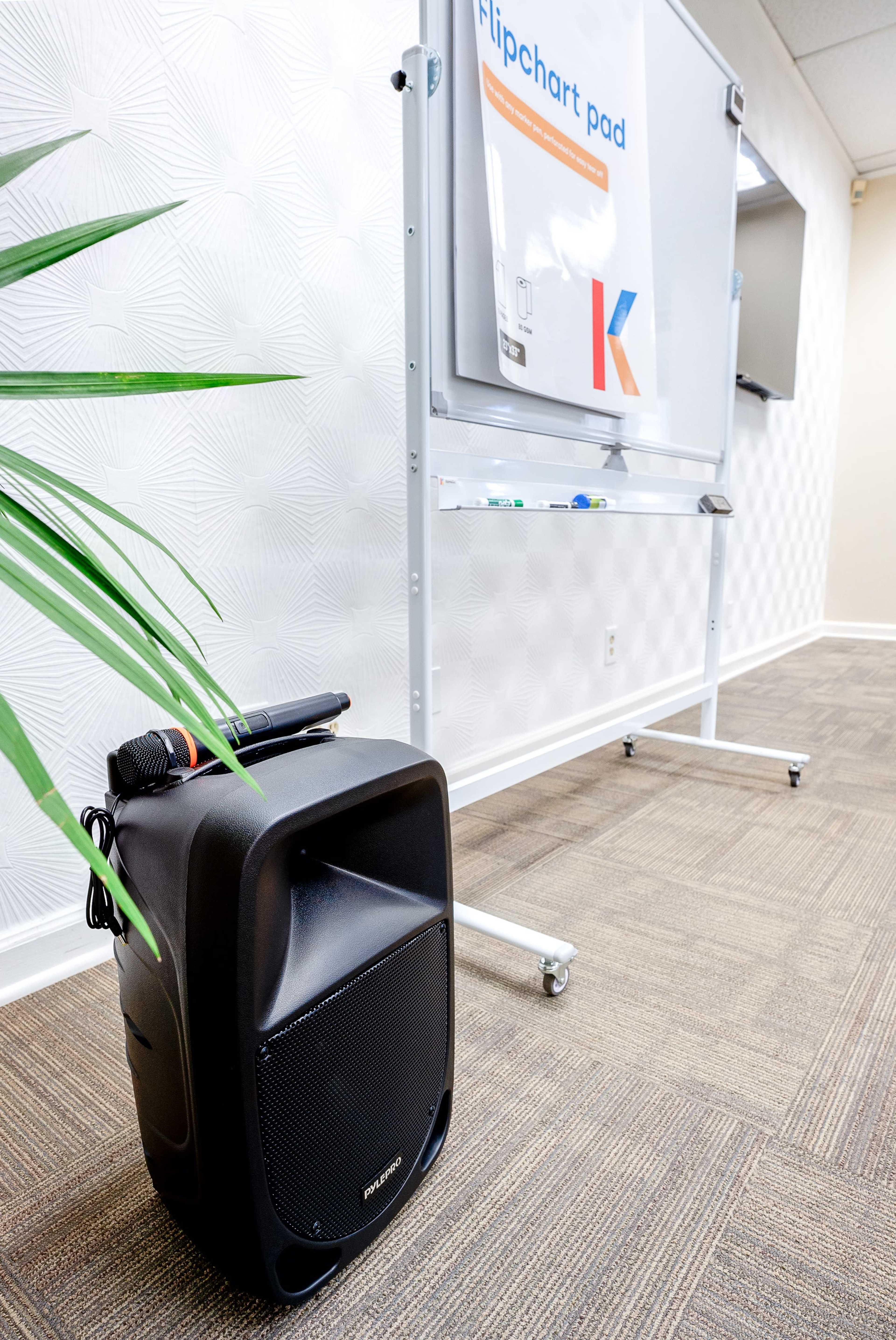 A portable speaker sits on the floor next to a whiteboard with a clipchart pad in a professional setting.