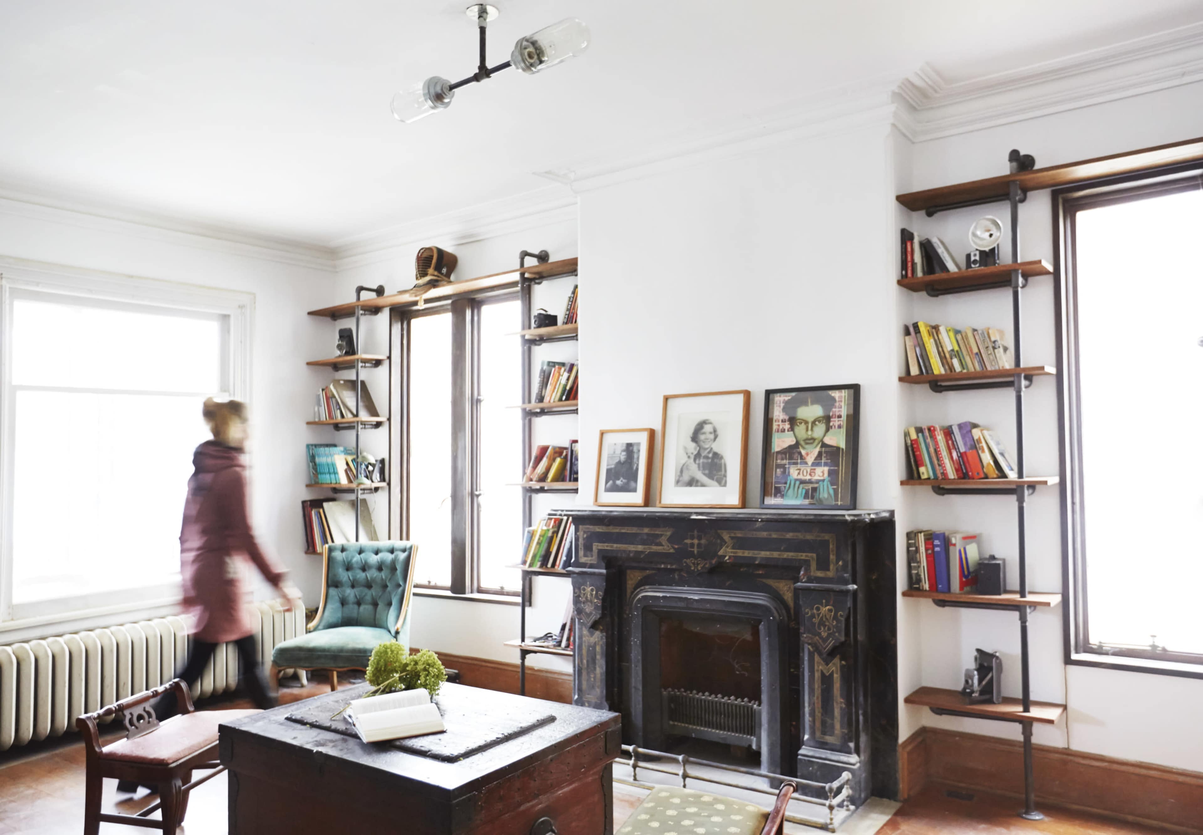 A woman walks past a bookshelf and a fireplace in a well-lit room with large windows.