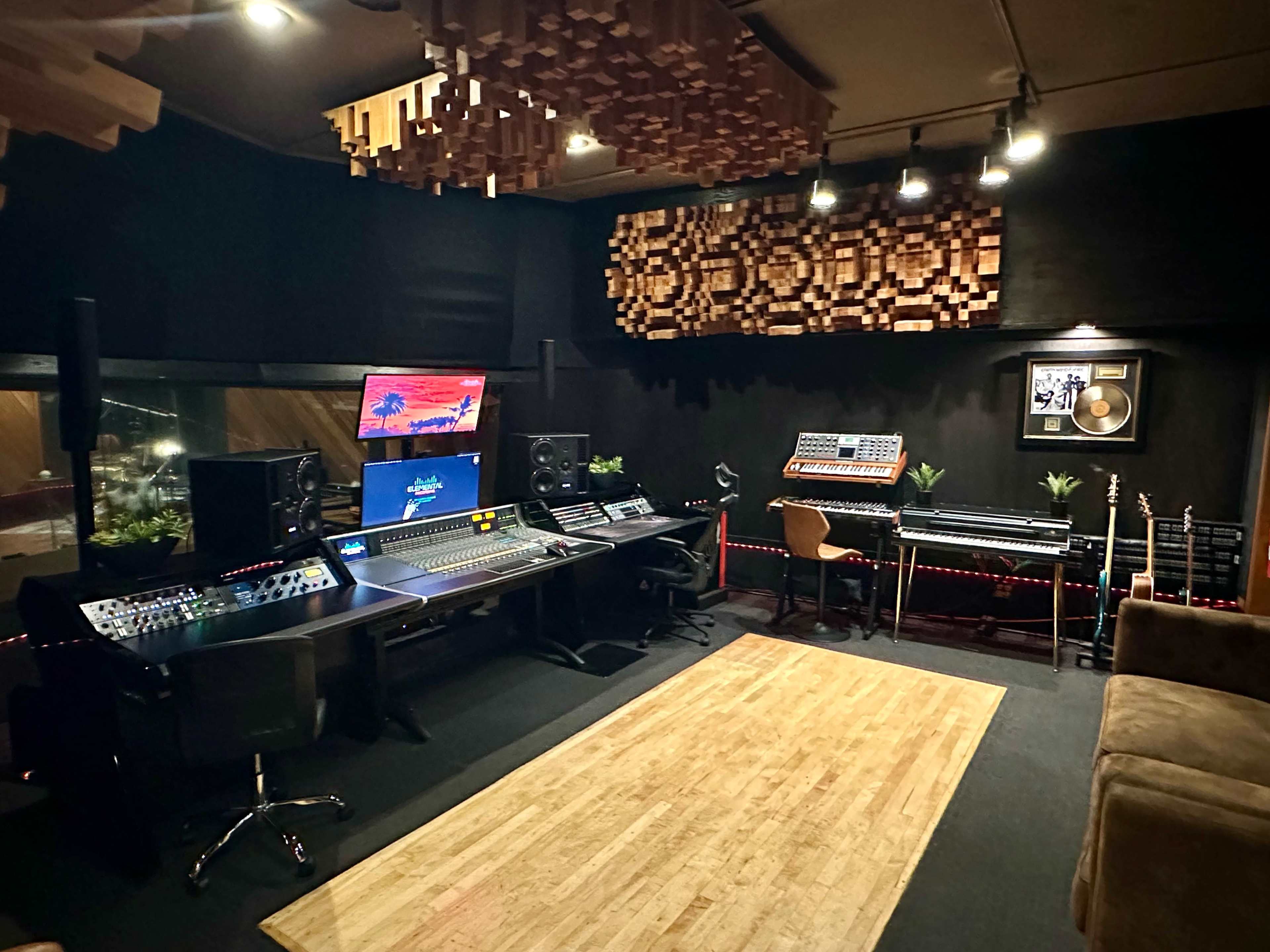 The image shows an interior view of a music recording studio featuring a large mixing console, monitors, synthesizers, and a wooden floor with a cozy seating area.