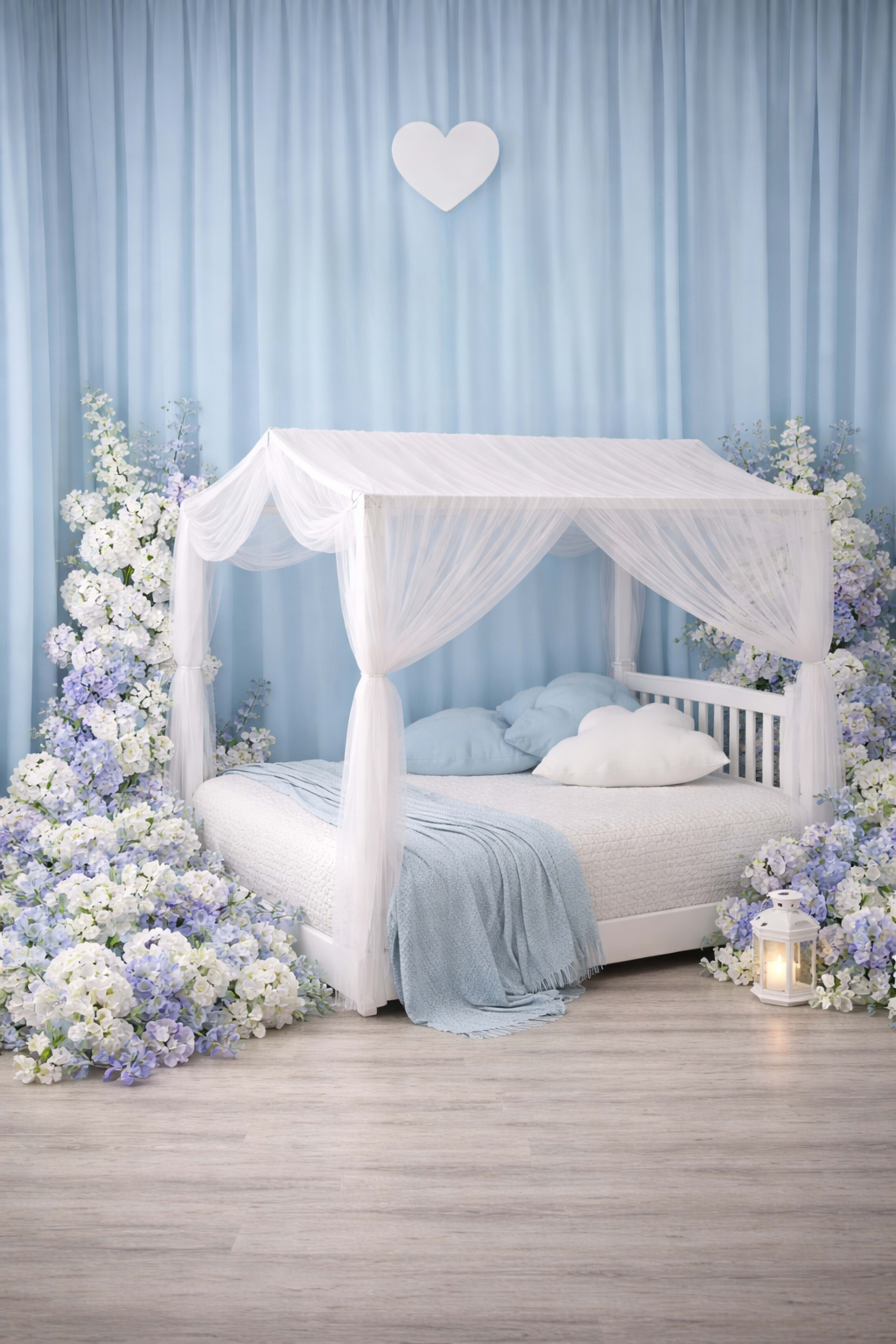 A white canopy bed is surrounded by lush arrangements of blue and white flowers, with a light blue backdrop and wooden flooring.