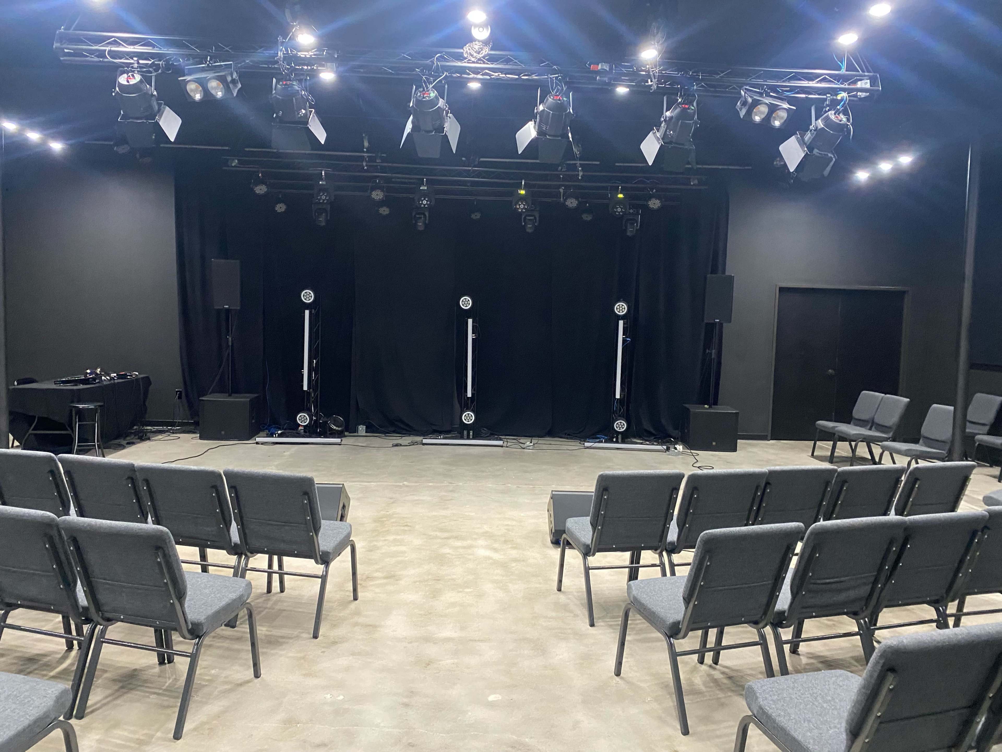 A staged performance area with a black backdrop, rows of gray chairs, and several lights mounted overhead.