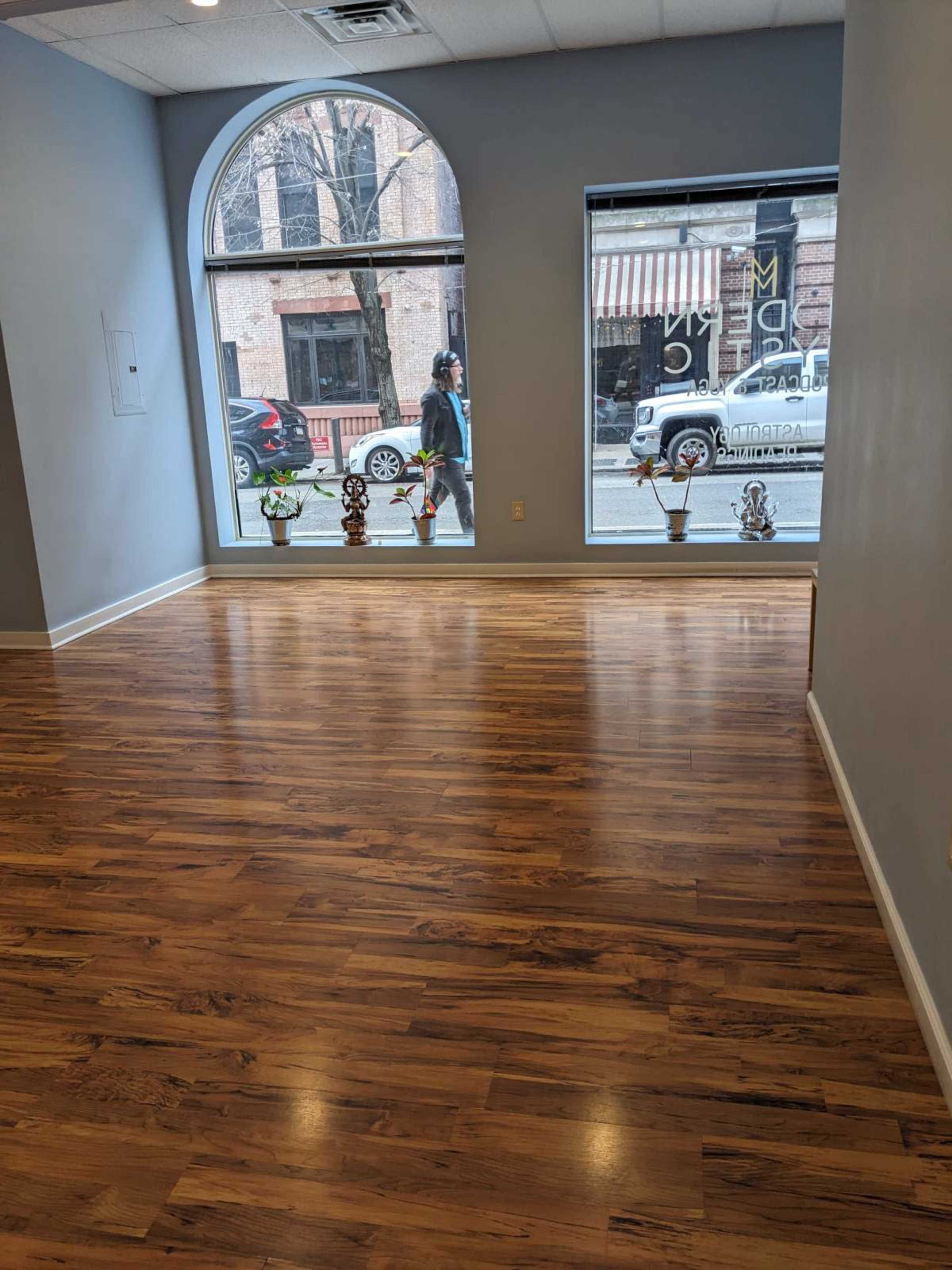 The image shows a spacious, empty room with hardwood floors and large windows facing a street, where a person is walking by.