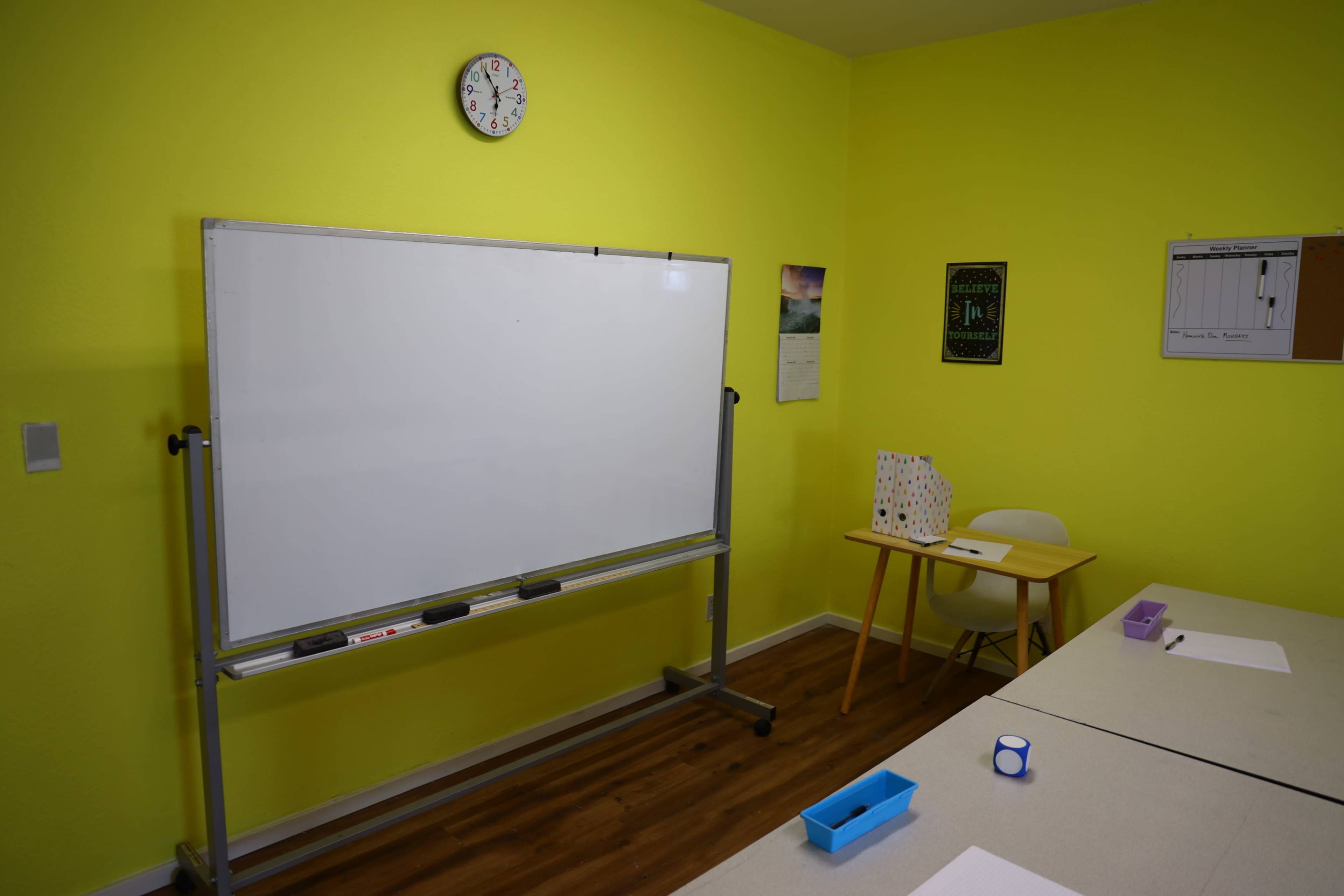 A bright room features a whiteboard on wheels, a clock on the wall, a calendar, and a table with supplies, all against a vibrant green background.