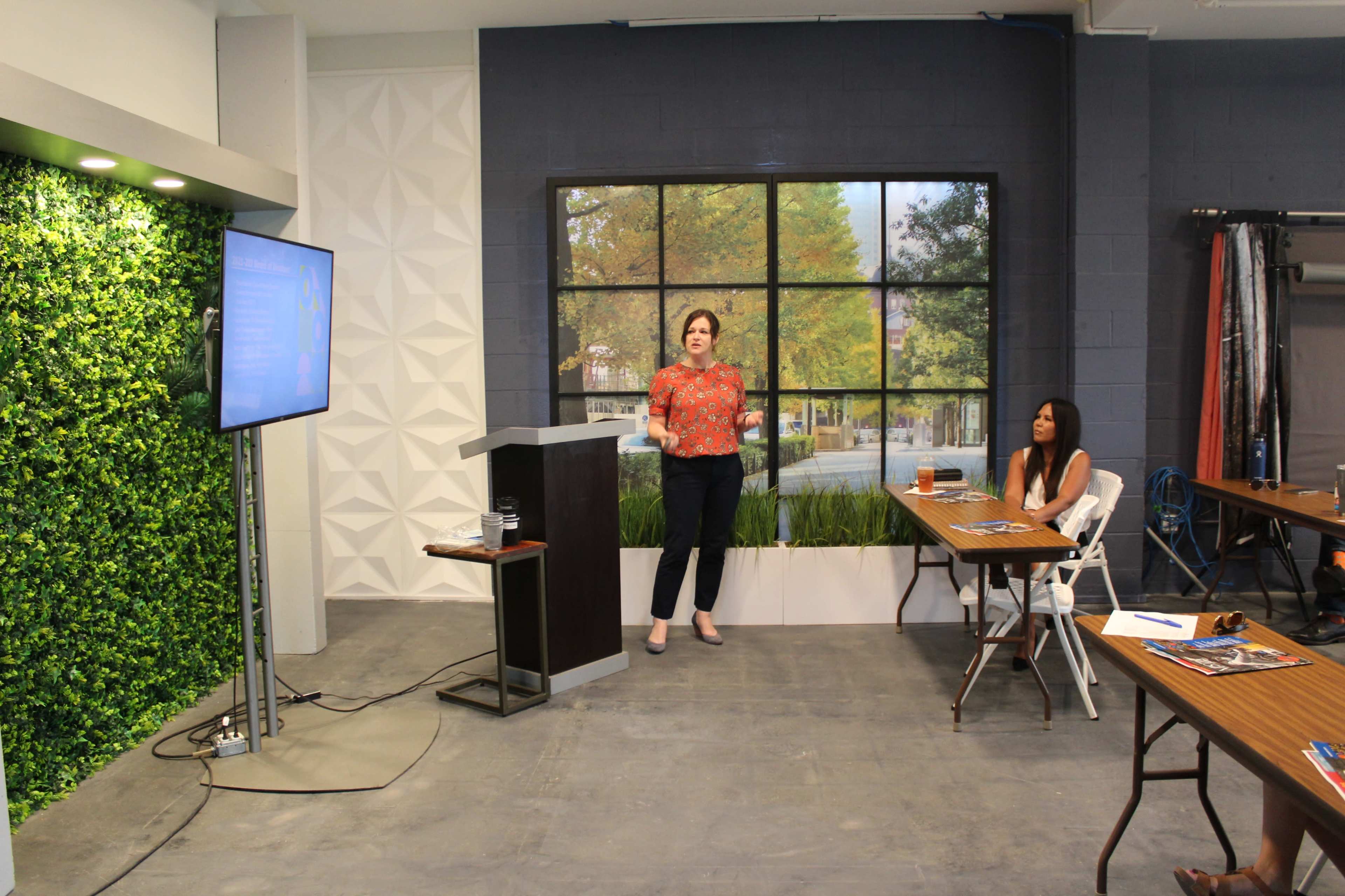 A woman presents at a podium in a well-lit room with greenery and a large window backdrop, while an audience member sits at a table.