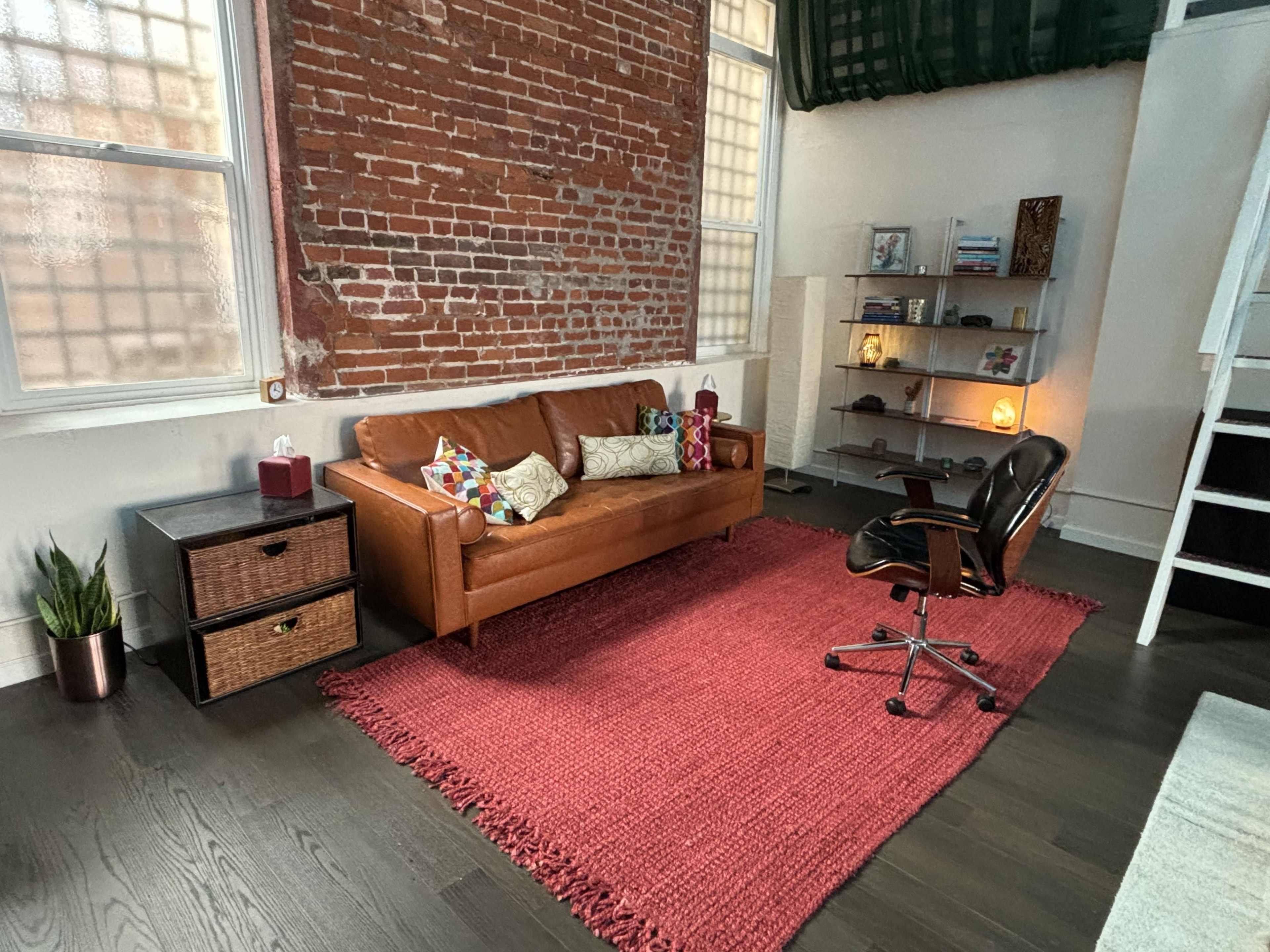The room features a brown leather sofa, a red area rug, a black office chair, and a bookshelf against a wall of exposed brick.