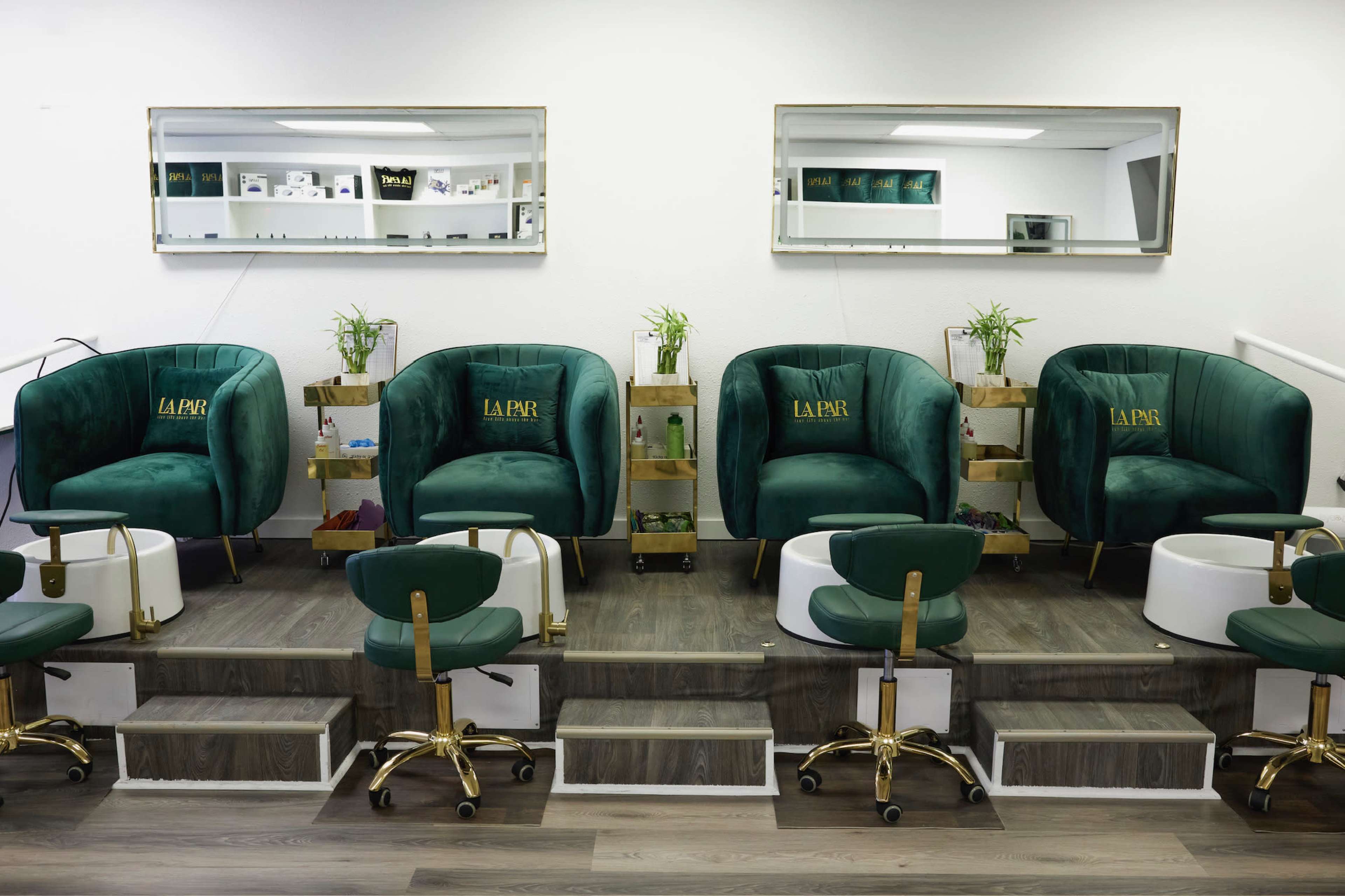 The image shows a modern nail salon featuring three green upholstered pedicure chairs, each with a complimentary white foot basin, situated in front of mirrors on a white wall.