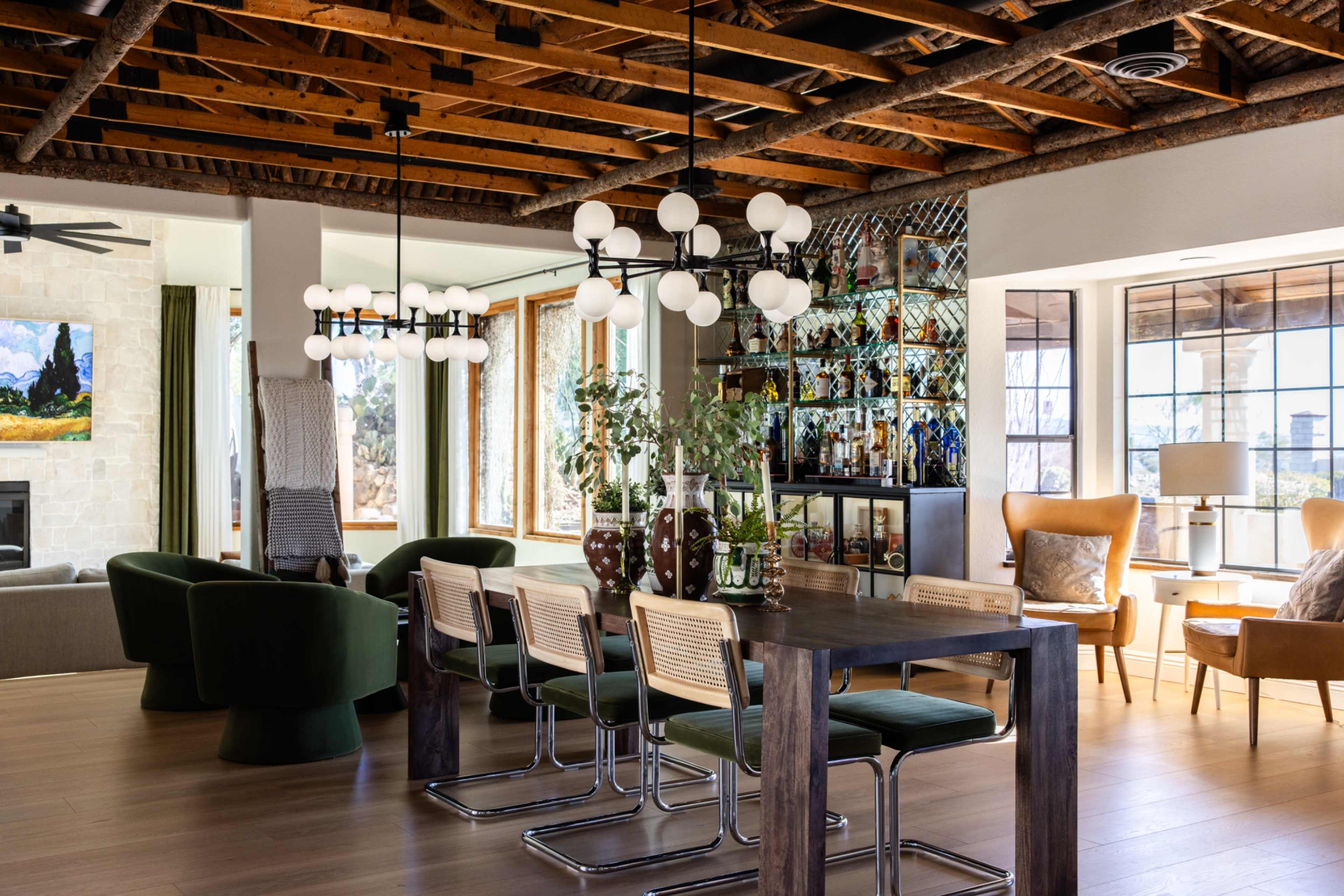 The image shows a modern living space with a dining table, green upholstered chairs, a bar area filled with plants and decorative items, and a large window overlooking an outdoor view.