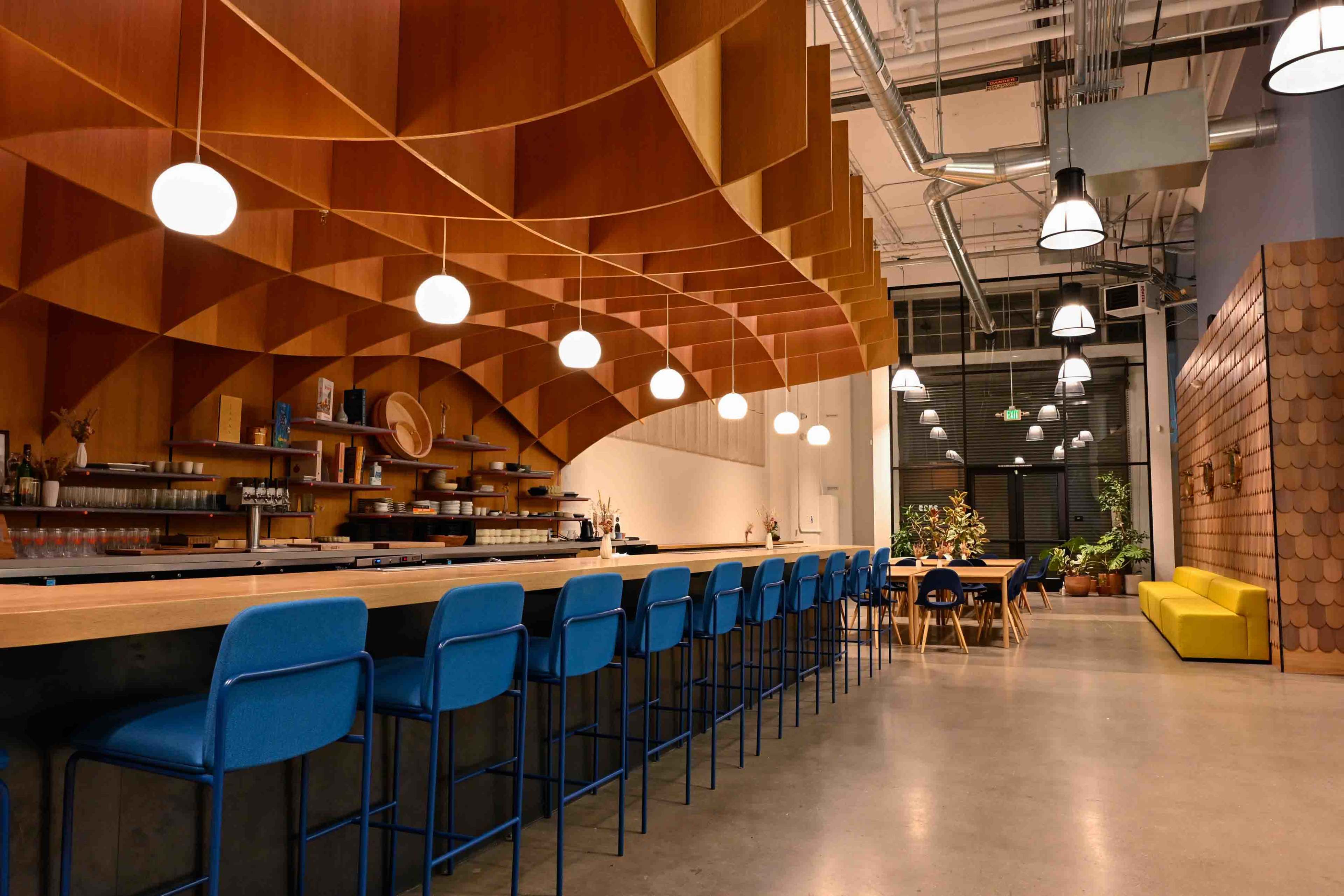A modern cafe features a curved wooden wall, a long bar with blue stools, and pendant lights hanging from the ceiling.