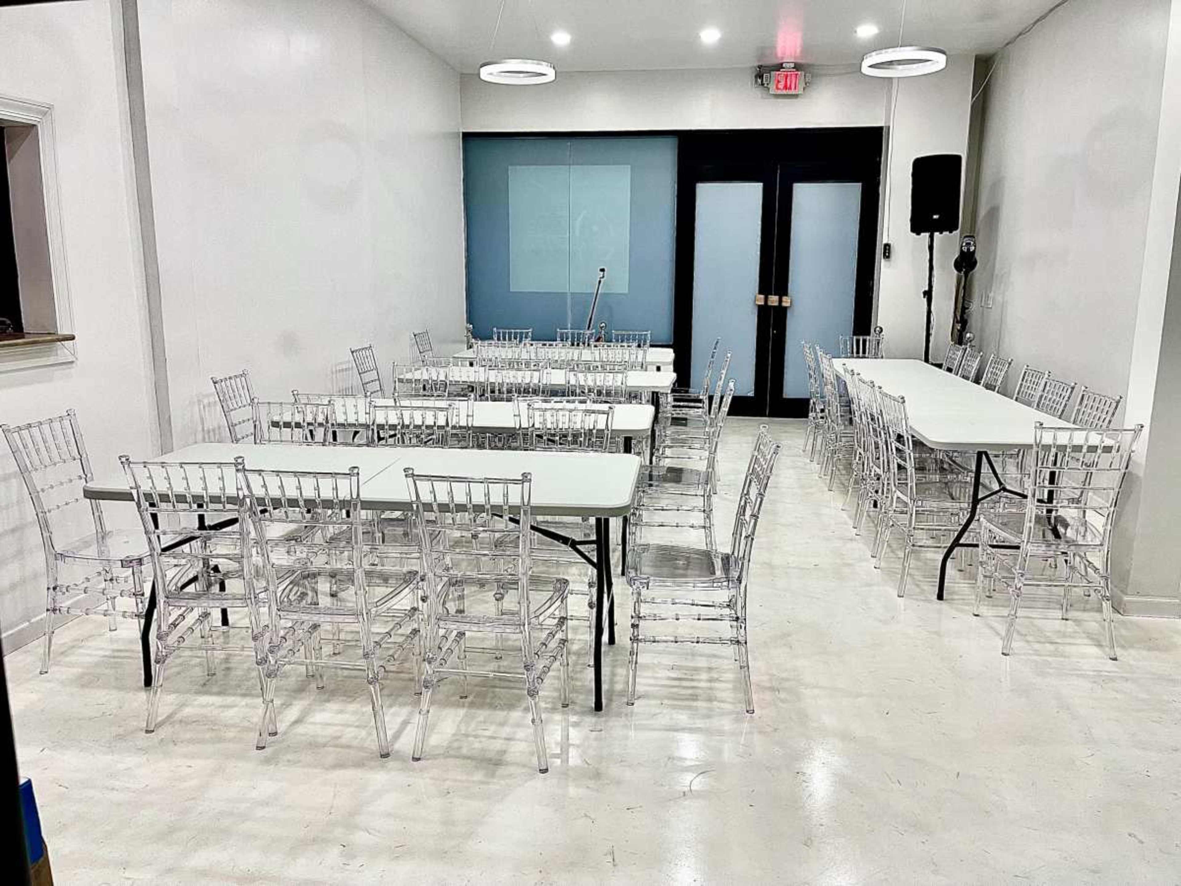 The image shows a white-walled room set up with multiple tables and clear chairs arranged for an event.