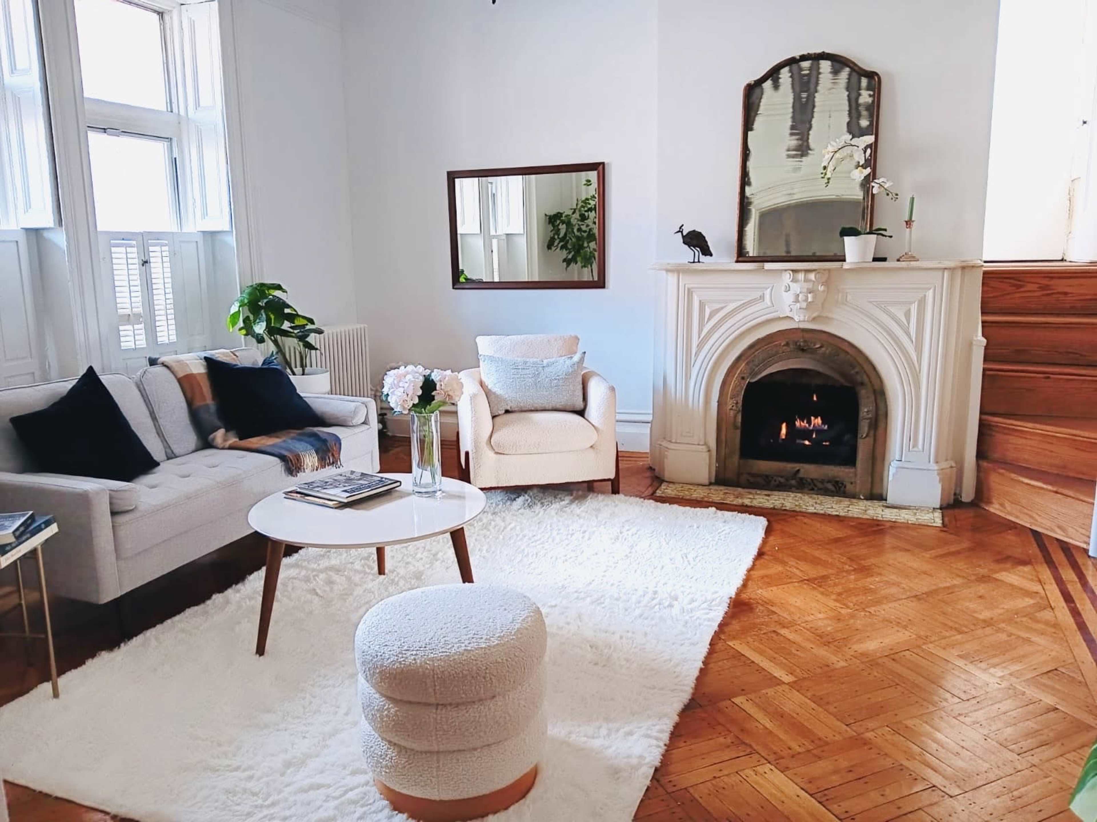 A cozy living room featuring a light-colored sofa, a small round coffee table, and a decorative fireplace with a mirror above it, all situated on a wooden floor with a large area rug.