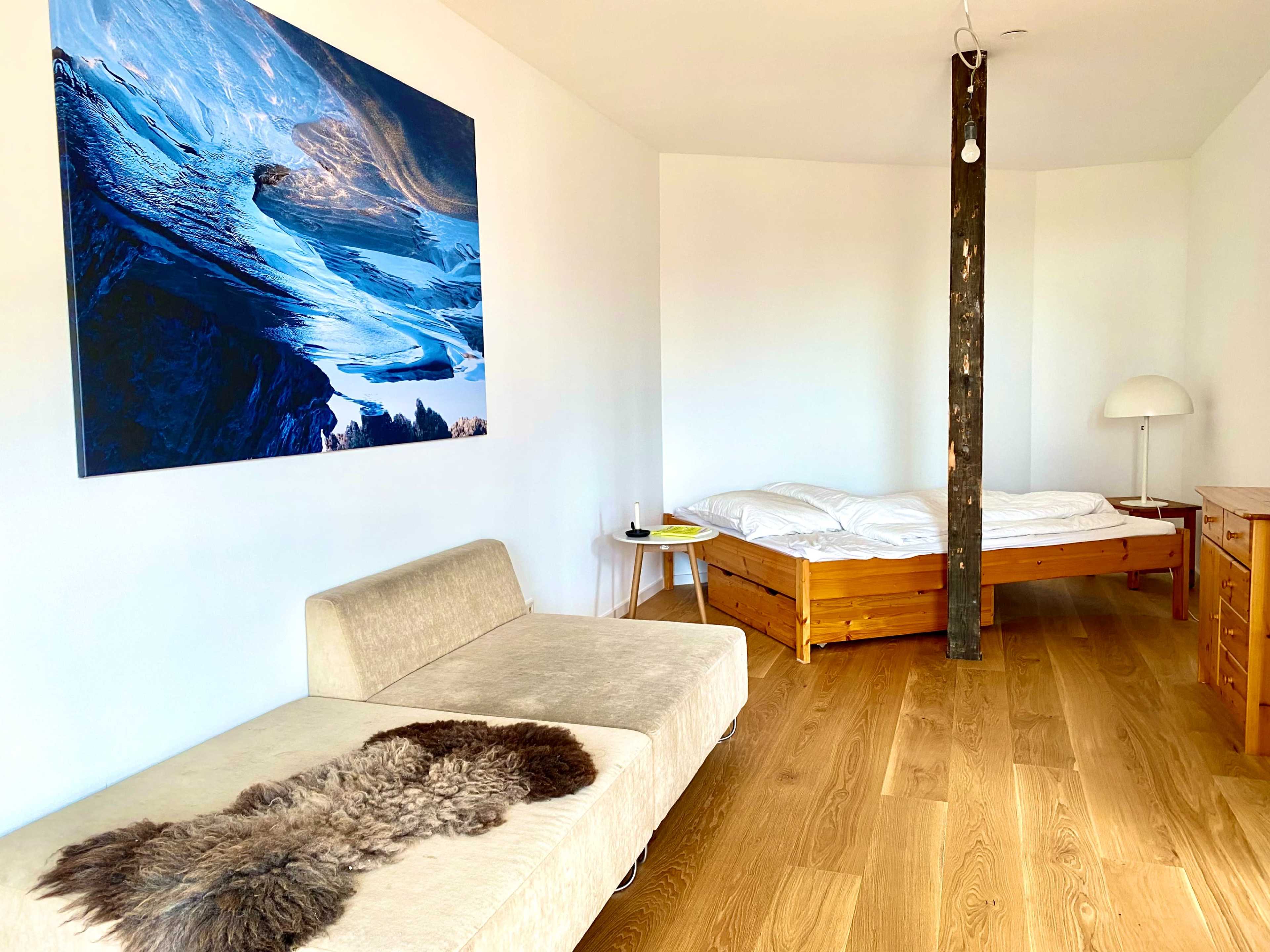 A minimalist room featuring a bed with white bedding, a wooden dresser, a gray sofa, and a large mountain landscape painting on the wall.