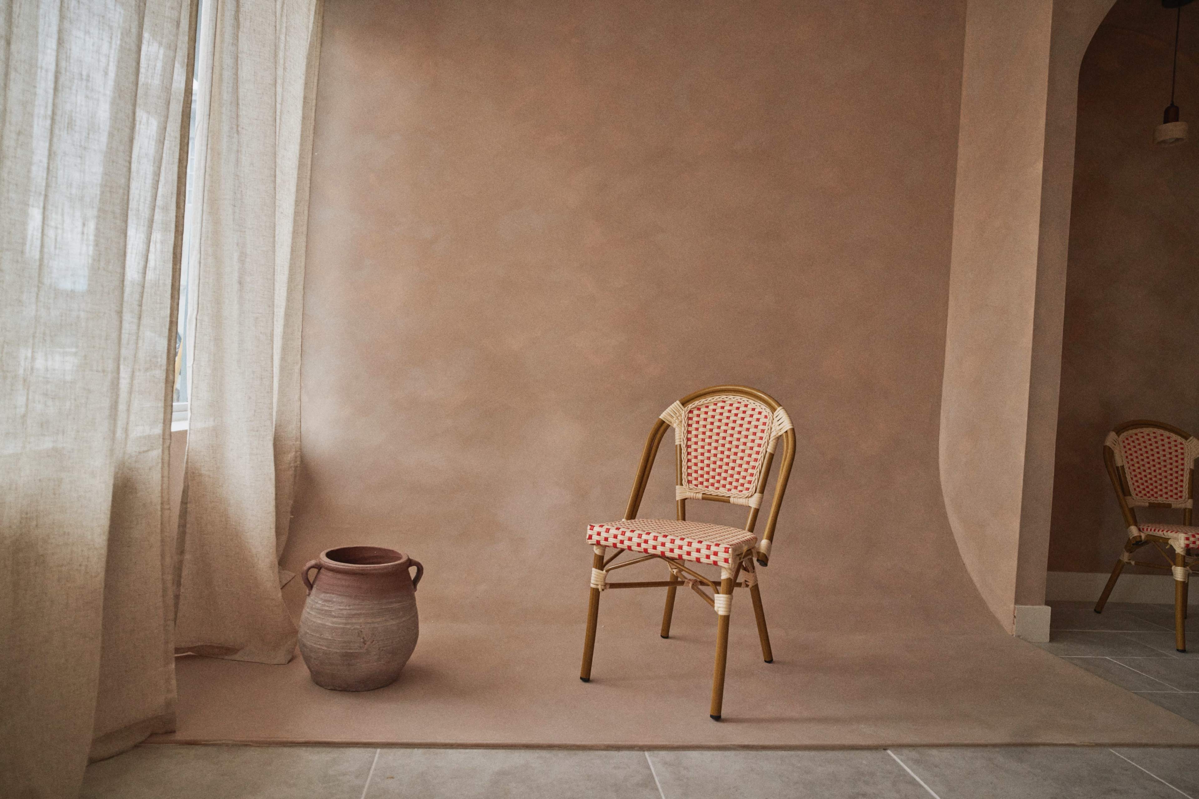 The image shows a minimalist interior scene featuring a patterned chair and a clay pot against a textured beige wall with a soft light filtering through sheer curtains.