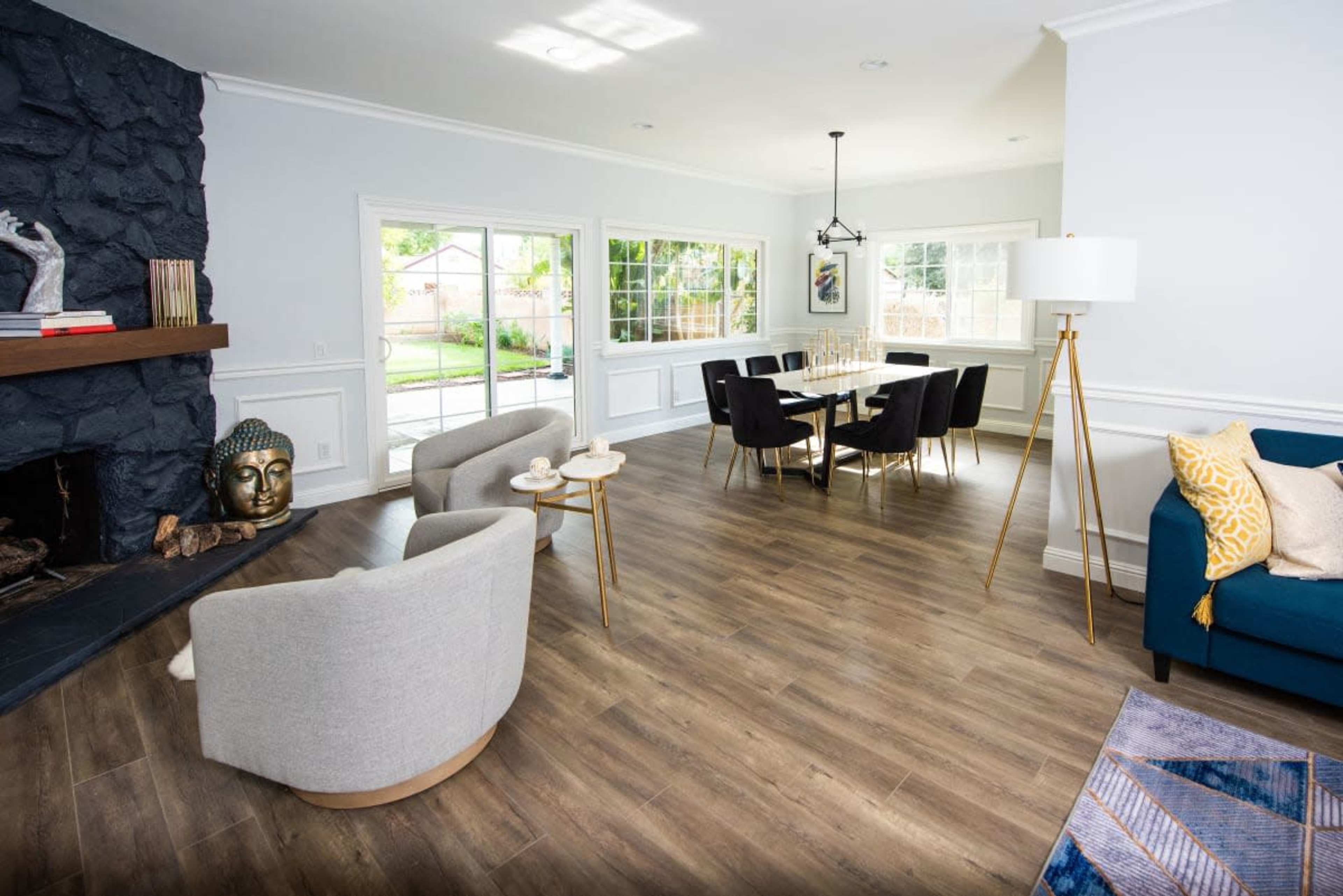 The image shows a modern living room and dining area featuring a stone fireplace, large windows, and a combination of stylish furniture, including a blue sofa and a dining table with chairs.